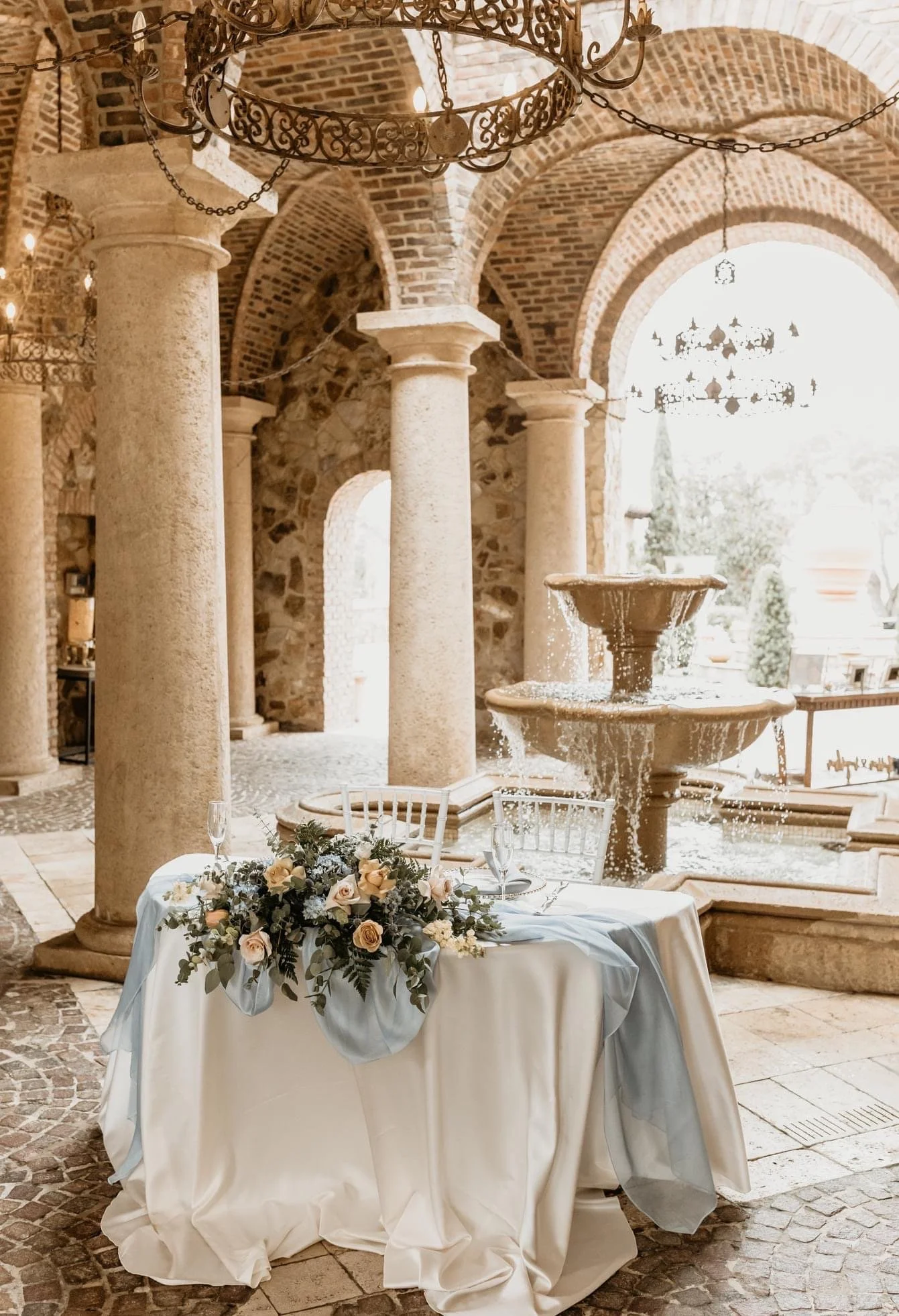 Elegant stone architecture with archways and columns, a decorative fountain, and a table adorned with white and light blue tablecloths, floral arrangement, and champagne glasses.