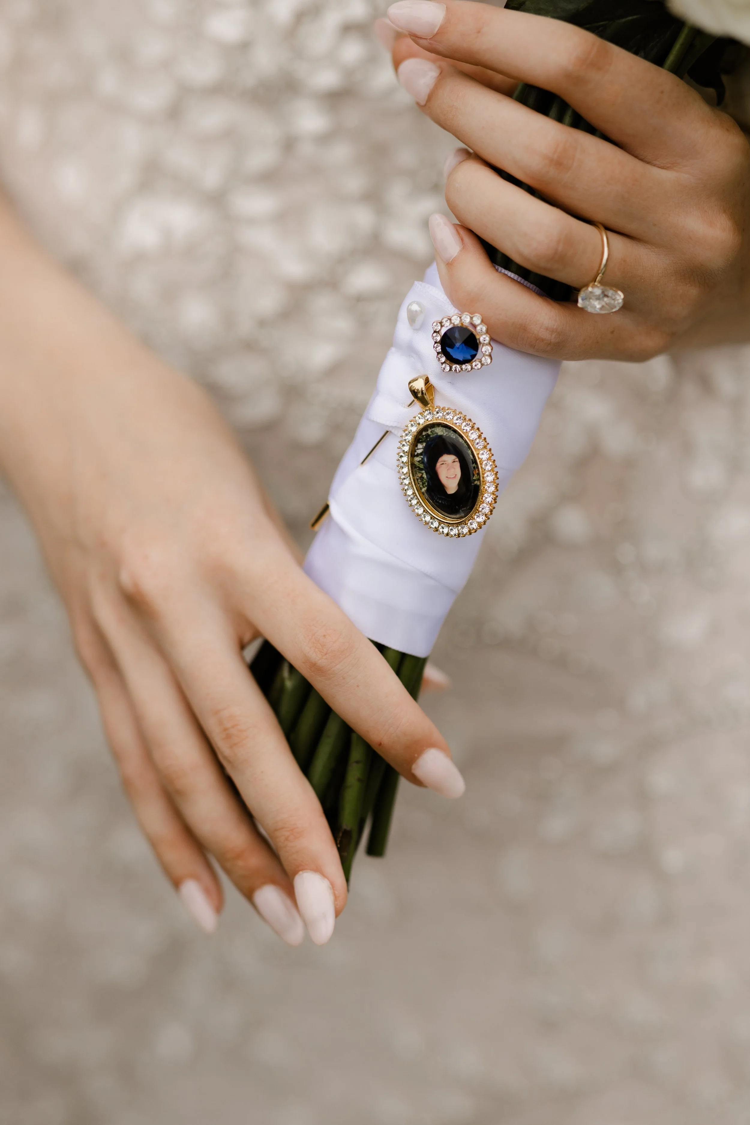 Close-up of a bride's hands holding a bouquet wrapped in white ribbon. The bouquet features a pin with a gemstone and a pendant with a photo. The bride has manicured nails and is wearing a ring.