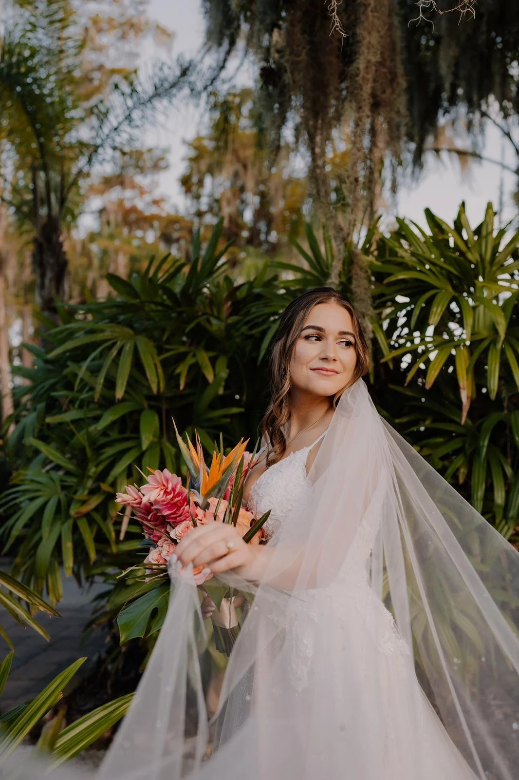 Bride in white dress holding a bouquet with tropical flowers, standing outdoors with lush green foliage in the background.