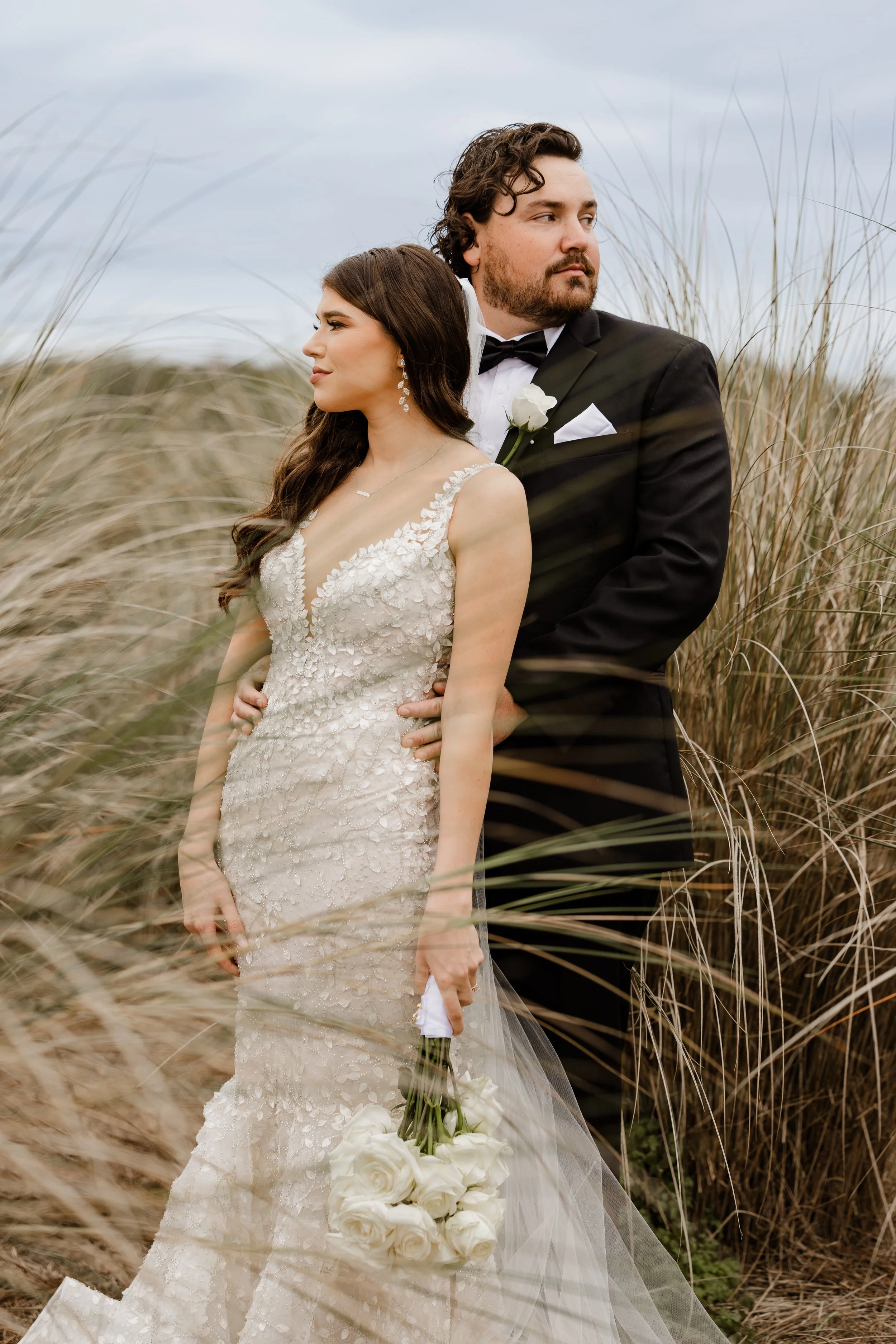 A bride and groom standing in tall grass, with the bride in a white lace wedding dress holding white roses and the groom in a black tuxedo, looking into the distance.