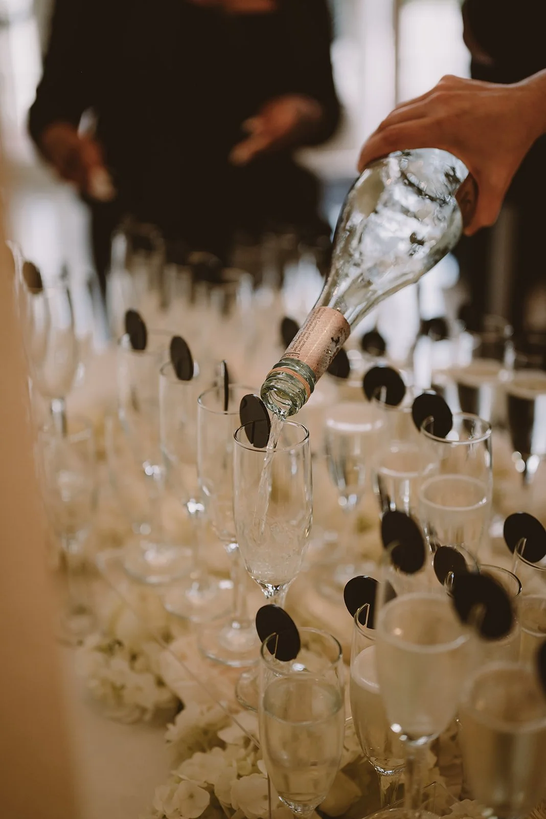 A person's hand pouring champagne into a row of tall glasses at an event, with decorative black round objects on the glass rims.