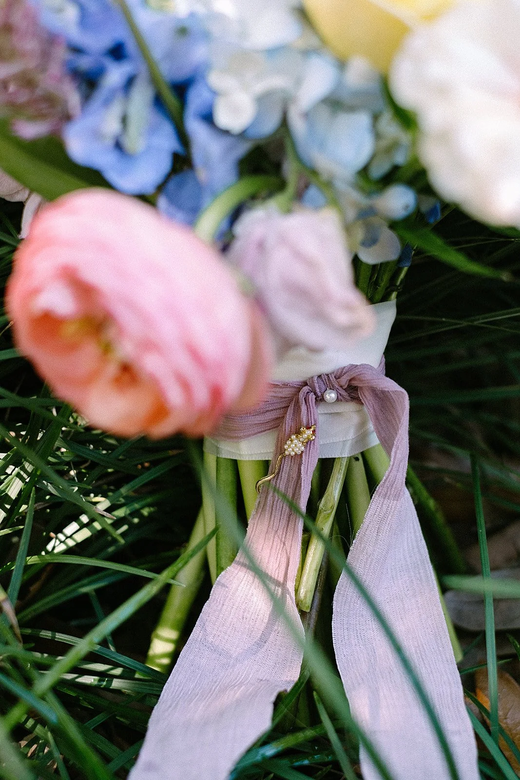 Close-up of a floral bouquet with pink, blue, and white flowers, wrapped with a light purple ribbon, featuring a small decorative pin.