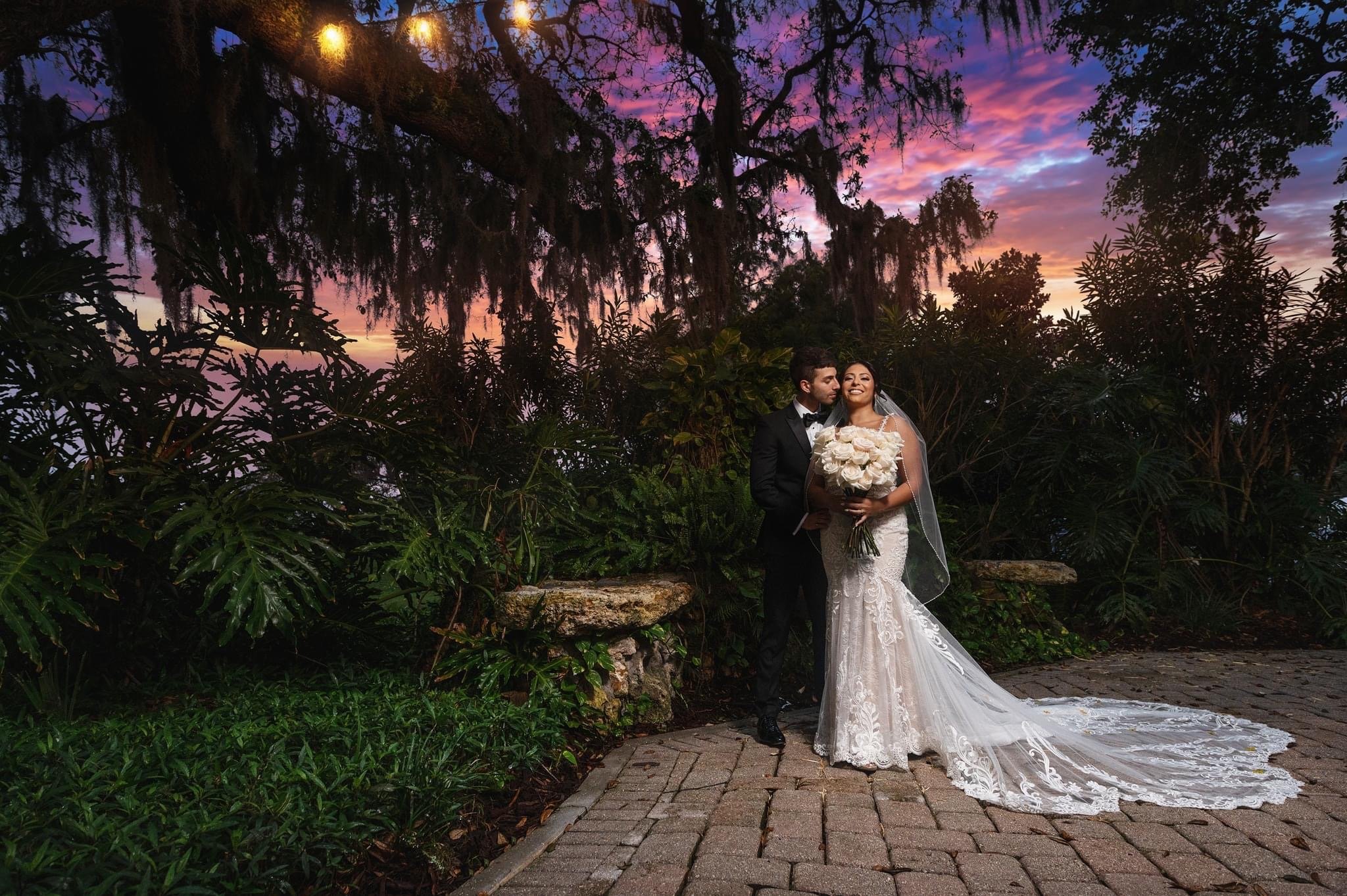 Bride and groom embracing in a garden at sunset, with hanging lights and lush greenery.
