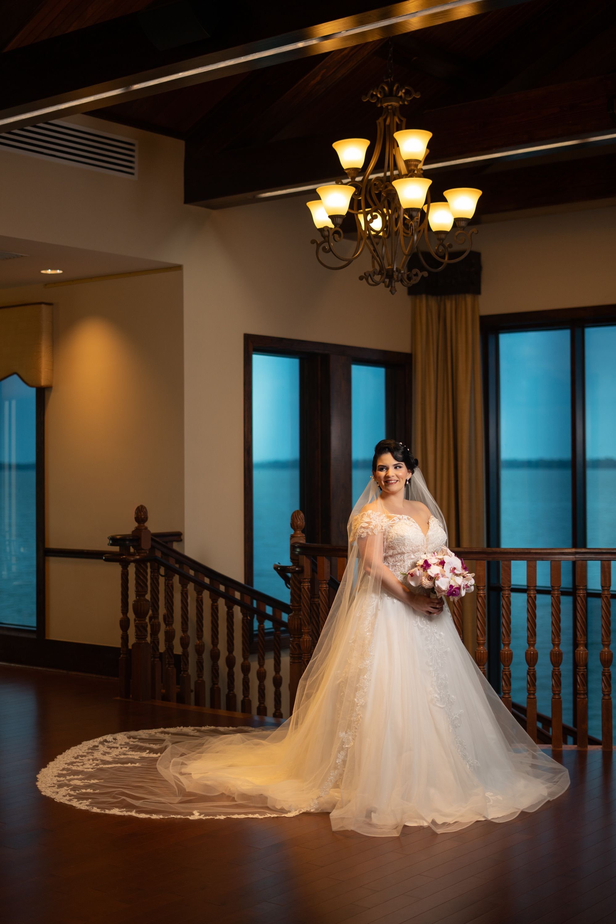 Bride in white wedding dress standing on wooden staircase with floral bouquet, chandelier above, large windows with view of water.