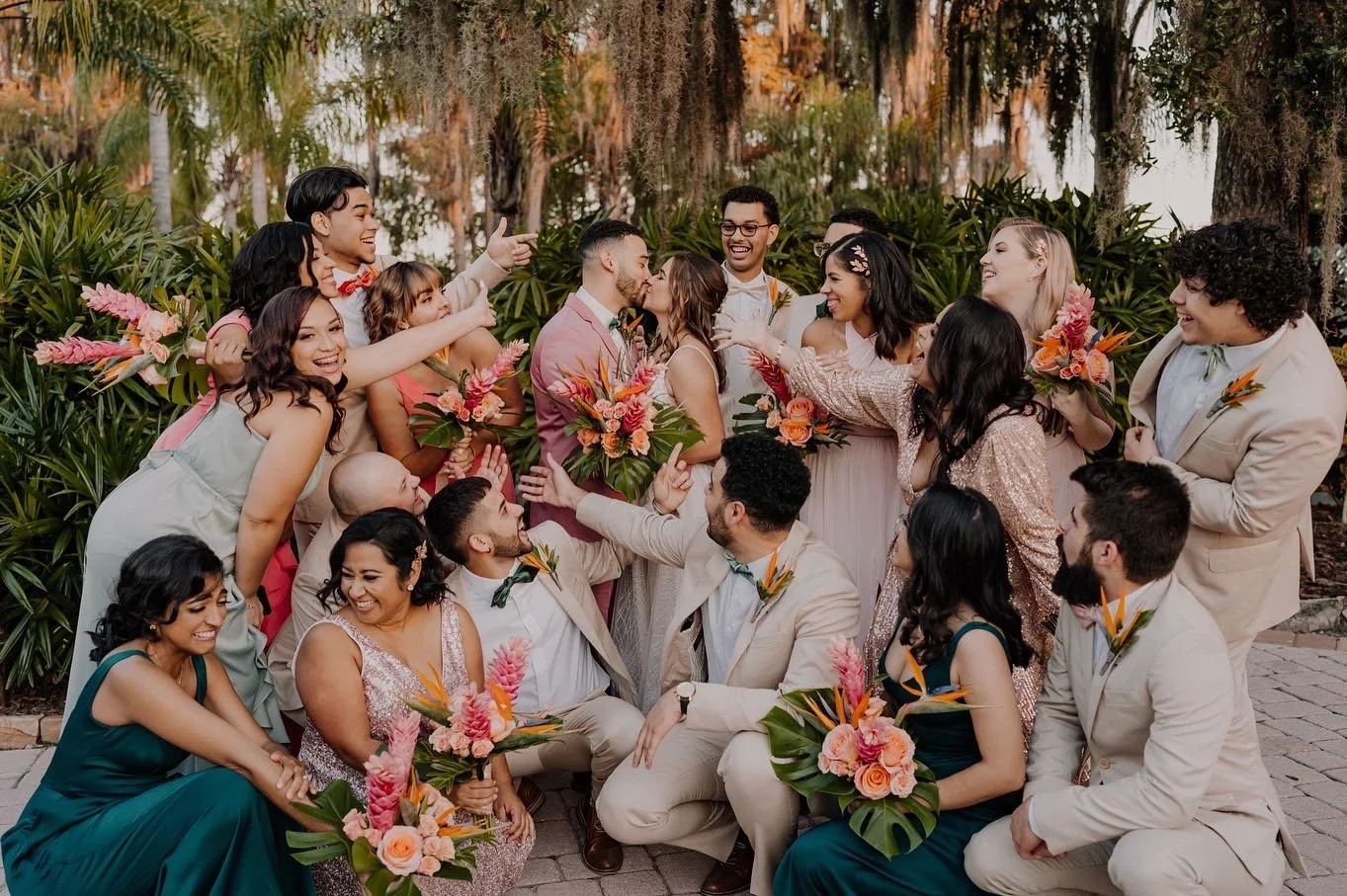 A wedding party gathered together outdoors with bridesmaids and groomsmen. They are dressed in formal attire, with women holding bouquets of pink and orange flowers. The couple, at the center, is kissing amidst joyful expressions and lively gestures 