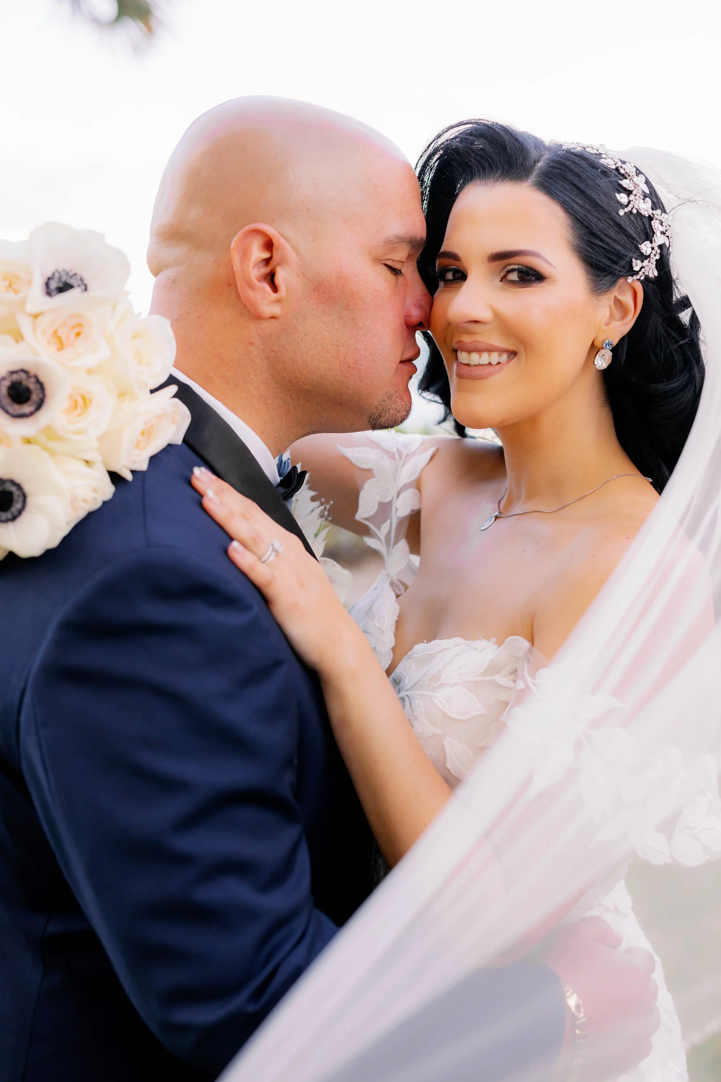 Close-up of a bride and groom embracing on their wedding day, with the bride smiling and wearing a floral lace dress and veil, and the groom in a suit with a white floral boutonniere.
