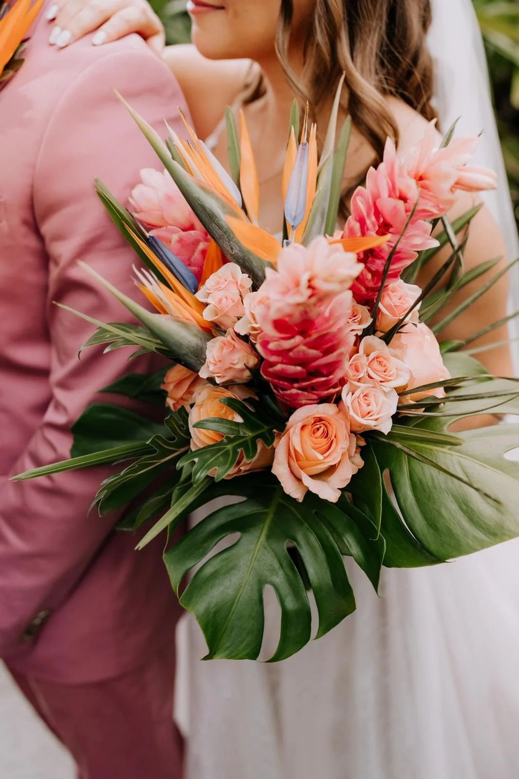 Bride holding tropical bouquet with pink and orange flowers, including roses and bird of paradise, alongside large green leaves.