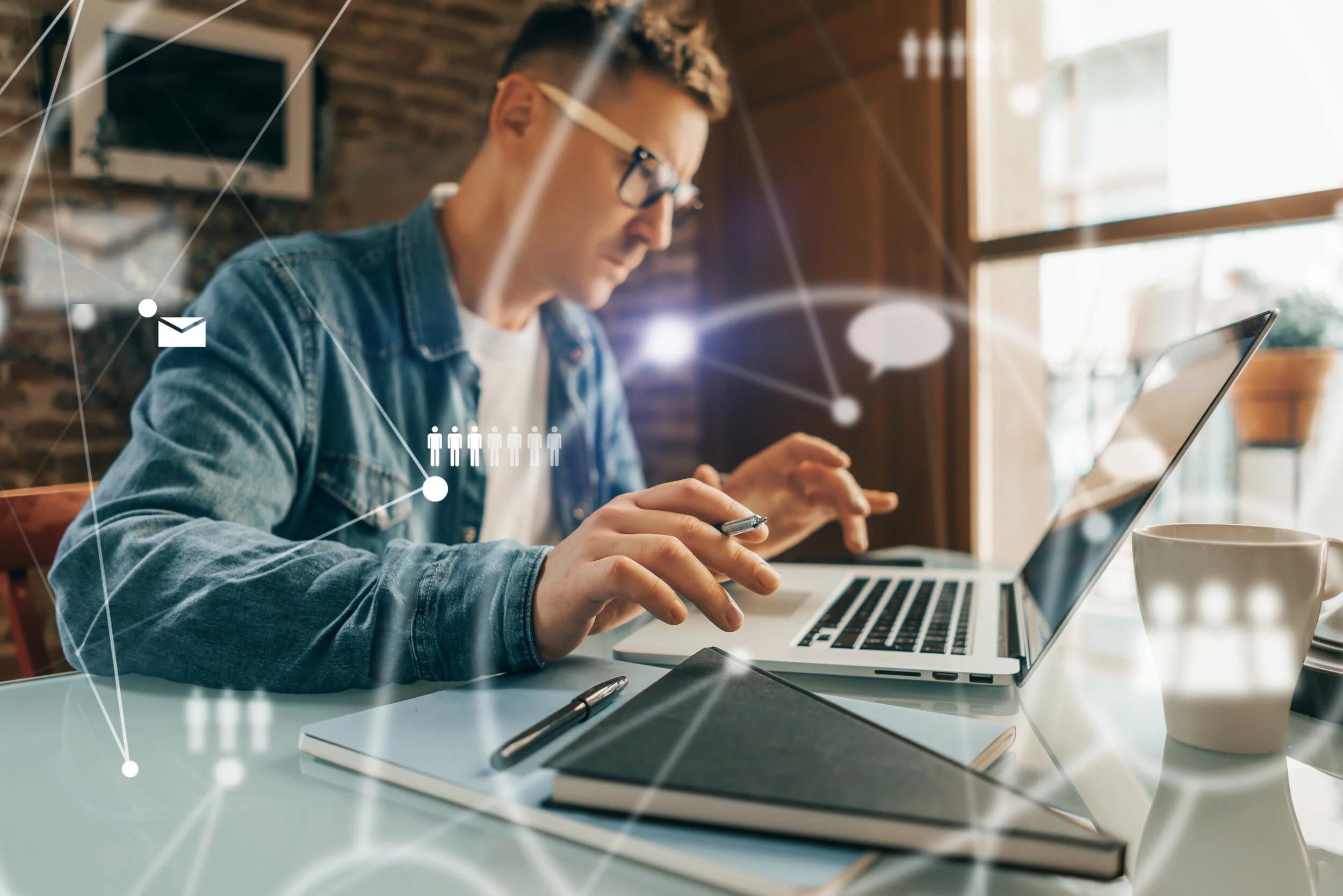 A young man with glasses working on a laptop at a table in a cozy cafe, surrounded by notebooks and a coffee mug, with digital icons and lines overlayed, indicating connectivity and communication.