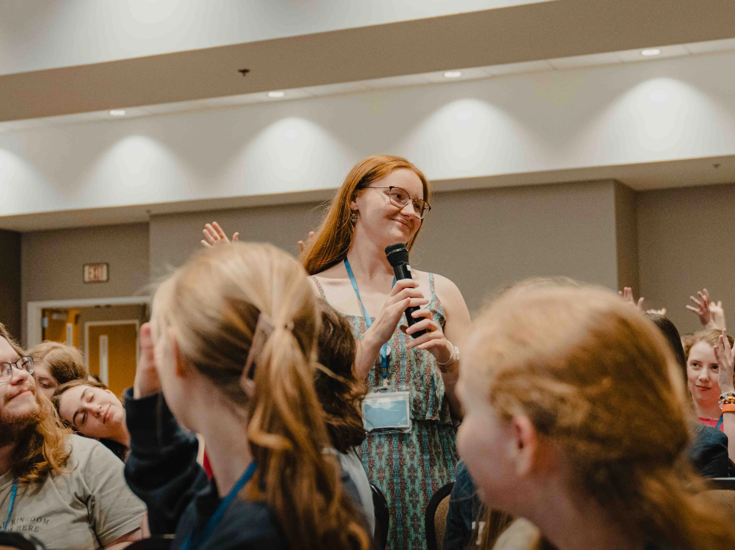 A woman with red hair, glasses, and wearing a patterned dress is speaking into a microphone at a conference or event, surrounded by a large group of people who are listening attentively.