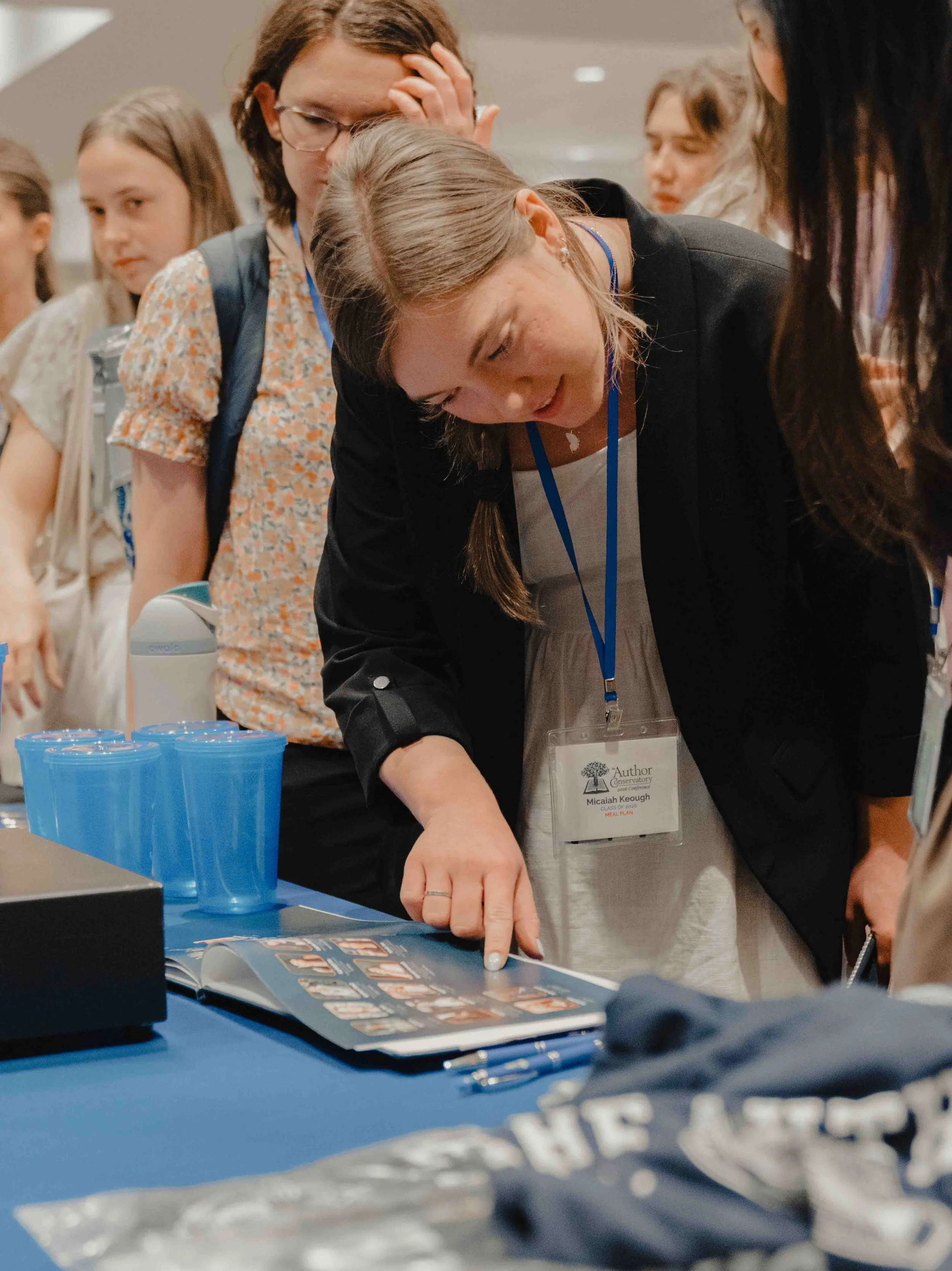 A group of people gather around a table at a conference or workshop, examining a catalog or brochure. One woman points at the page, and others look on. Items on the table include blue plastic cups, pens, and a brochure or catalog. The woman in the fo