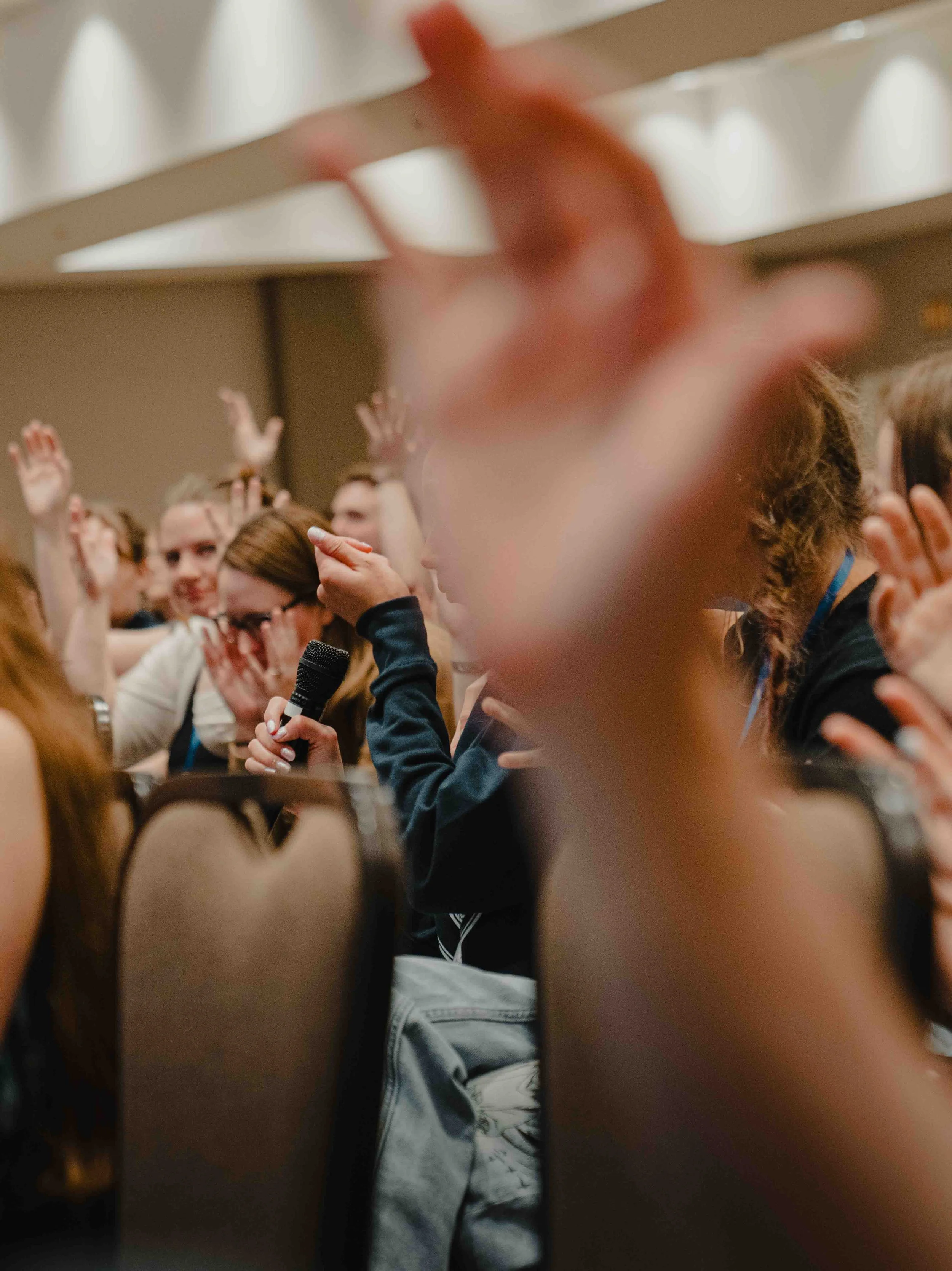 A person holding a microphone in a large audience, with several audience members raising their hands, and a person's arm and hand reaching toward the camera in the foreground.