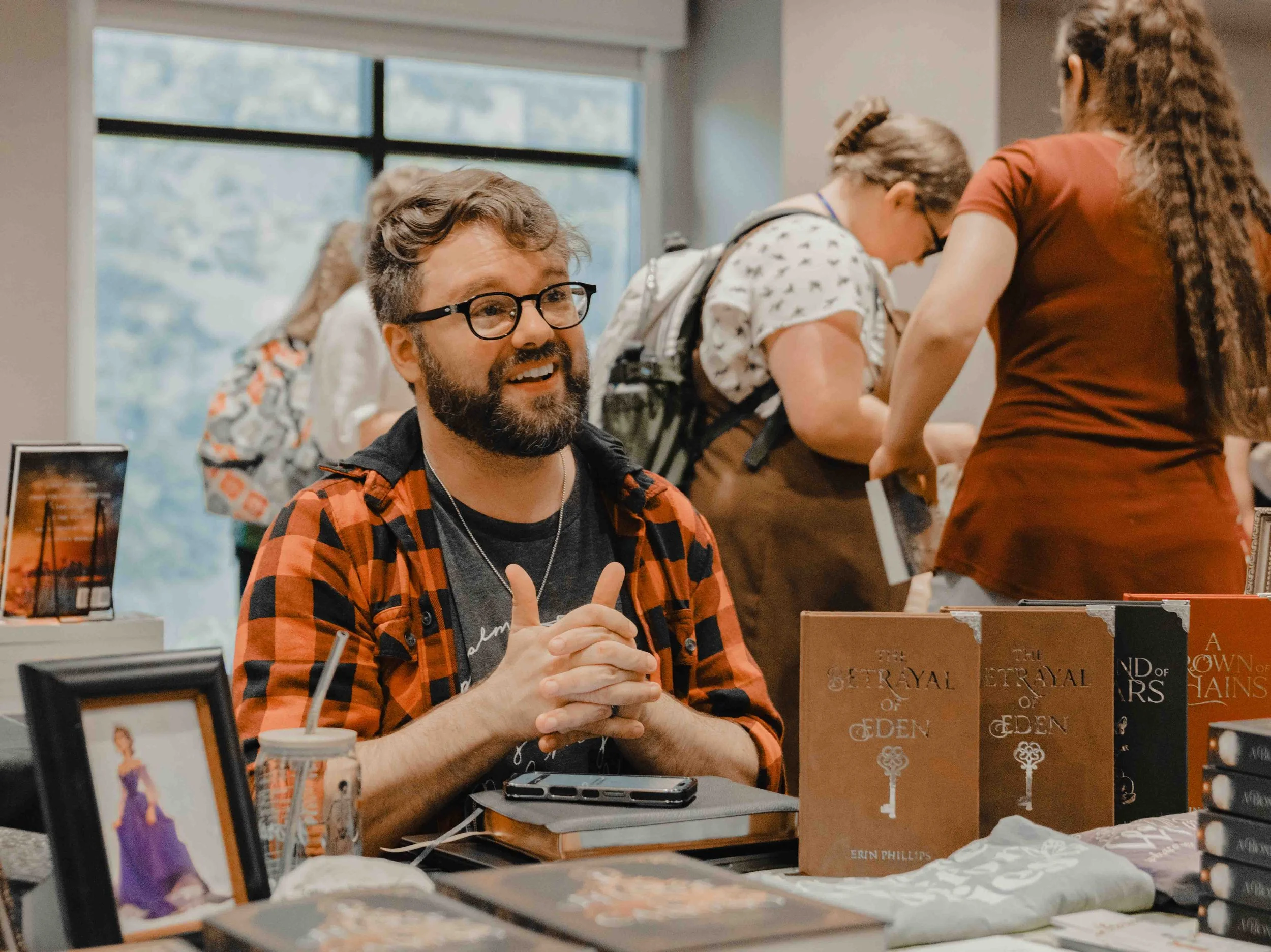 A man with glasses and a beard sitting at a table with books, bookshelves, and a woman with braided hair leaning over to talk to him at a book signing event.