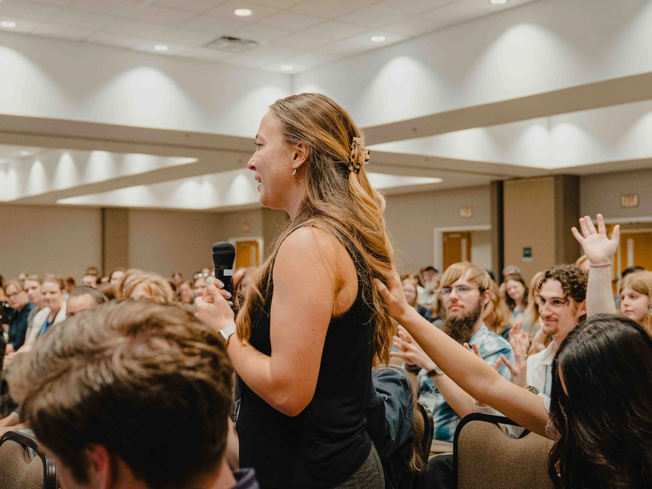A woman with brown wavy hair, wearing a black sleeveless dress, holding a microphone, standing among seated people in a large conference room with white ceiling lights.