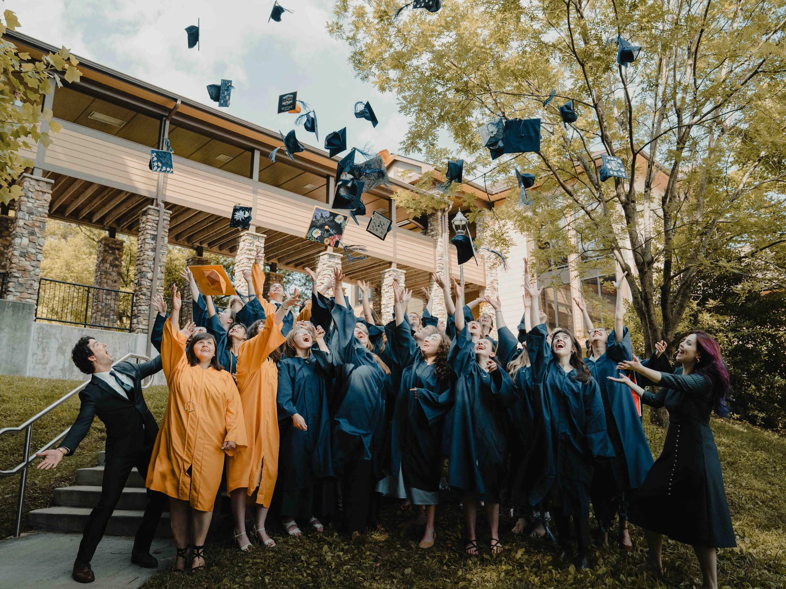 A group of graduates in caps and gowns celebrating outdoors, throwing caps into the air beneath a tree on a sunny day.