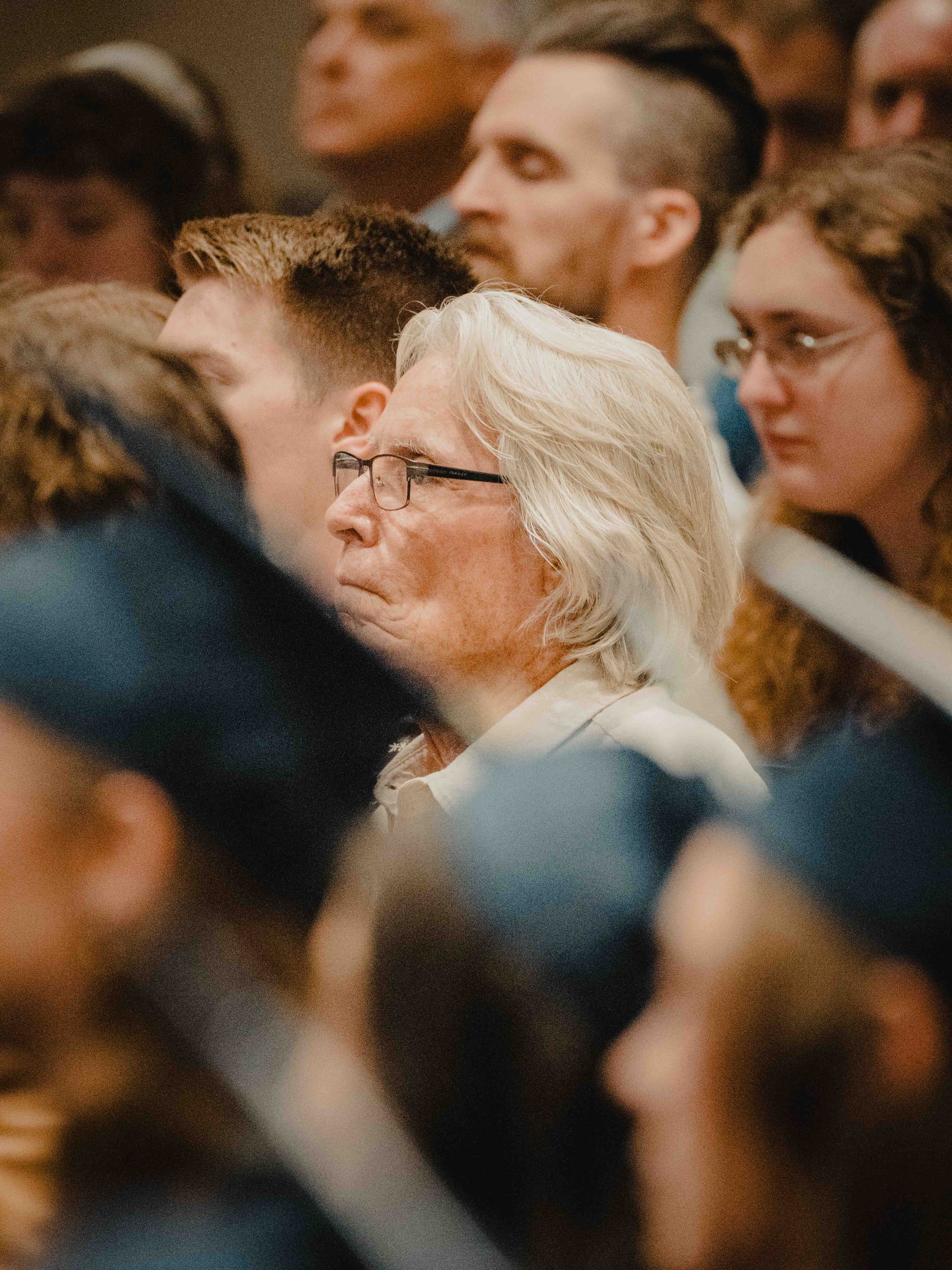 A group of diverse people in close proximity, facing in different directions, with various hairstyles and glasses, at what appears to be a crowded event or gathering.