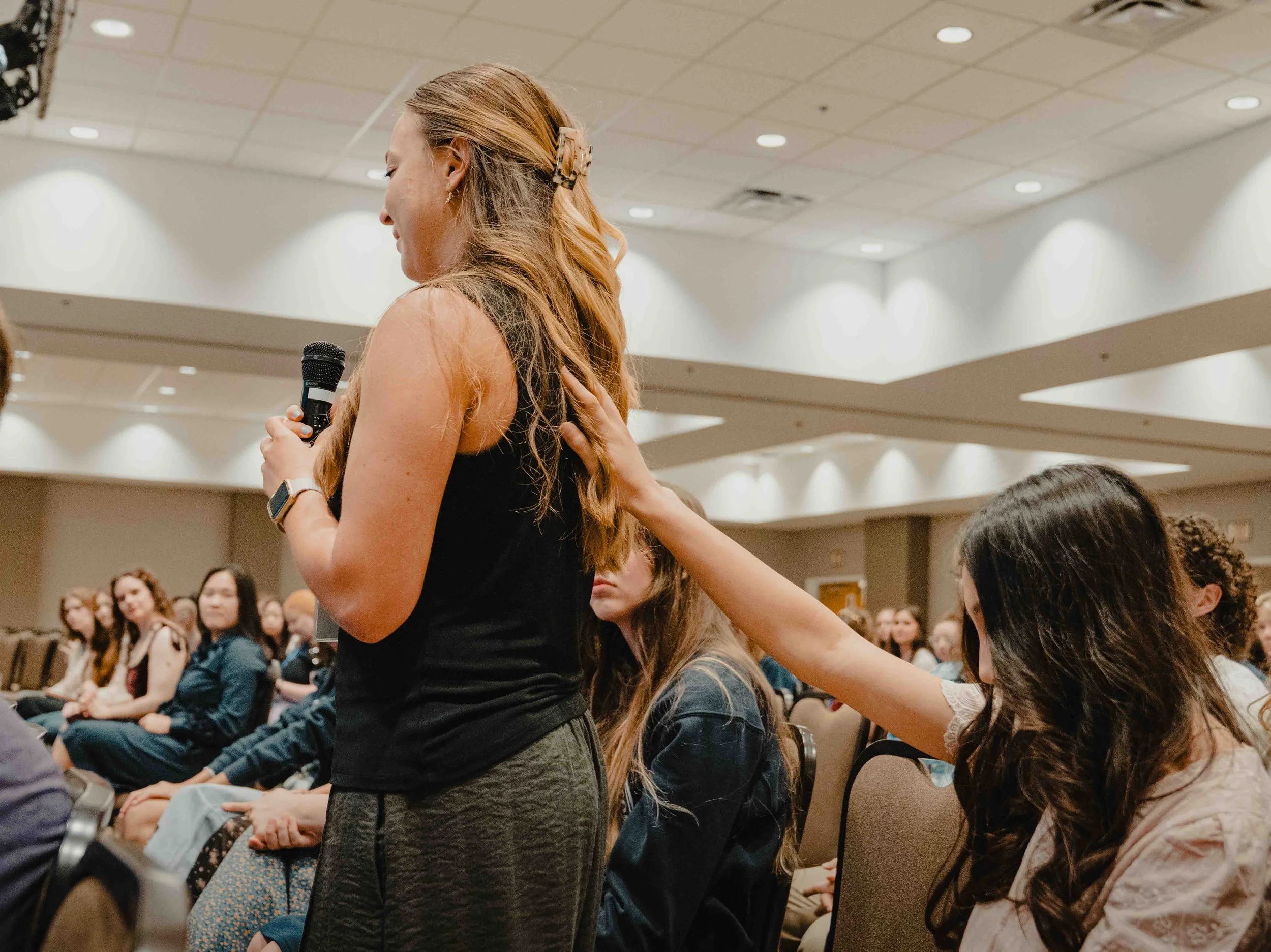 A woman with long red hair and a flower hair clip is holding a microphone and listening to a person in the audience. She is wearing a watch and a black top. The audience consists of seated women, some with curly hair, in a conference room with a susp