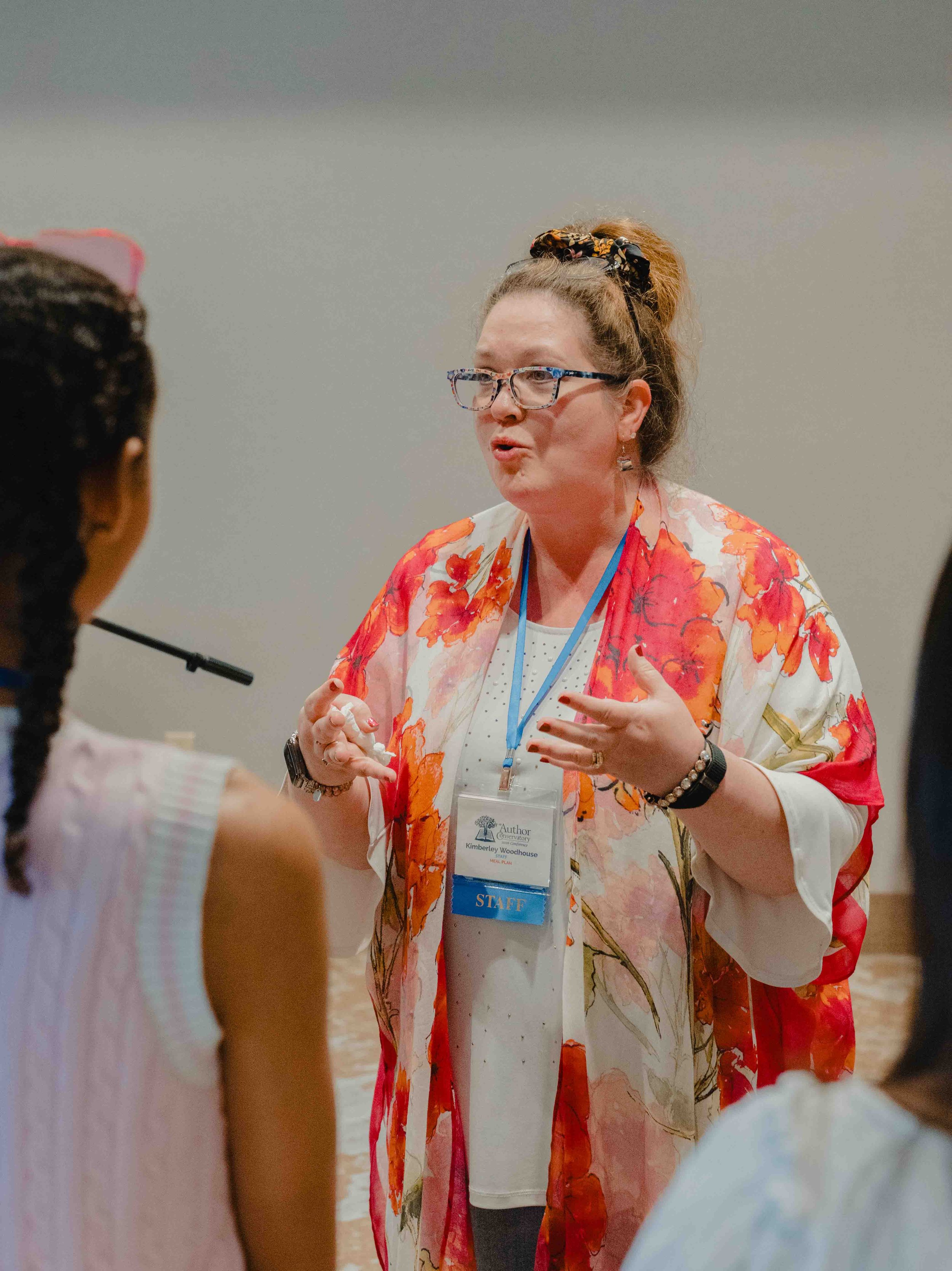 A woman with glasses and curly hair tied with a floral scrunchie is talking to a group of children. She is wearing a flower-patterned shawl and has a lanyard with a nametag that says 'Author' and 'Staff.' The children are in the foreground, with one 