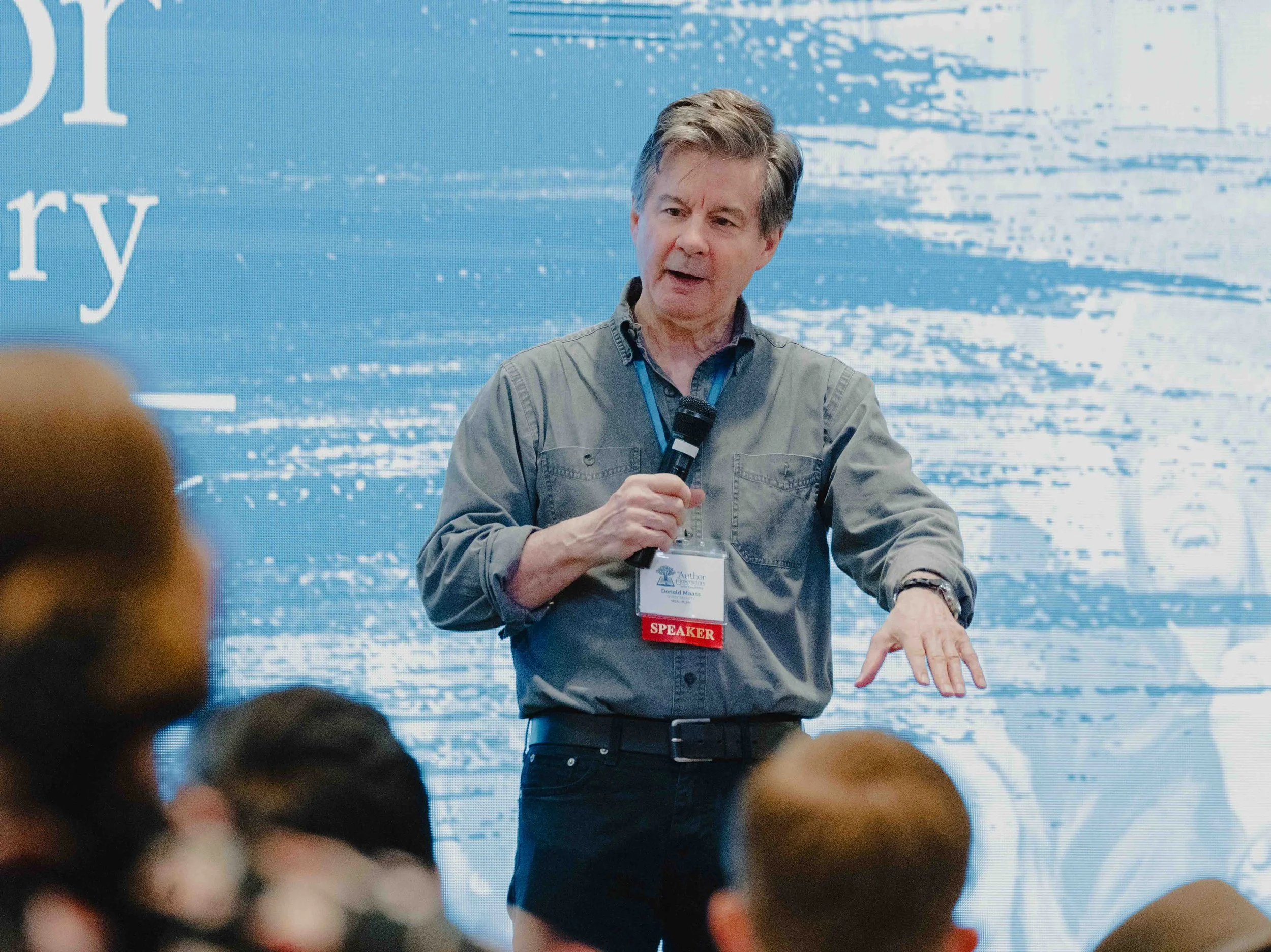 A man with gray hair, wearing a gray shirt with a 'SPEAKER' badge, speaking into a microphone at a conference or event with a large digital screen behind him displaying text and graphics.
