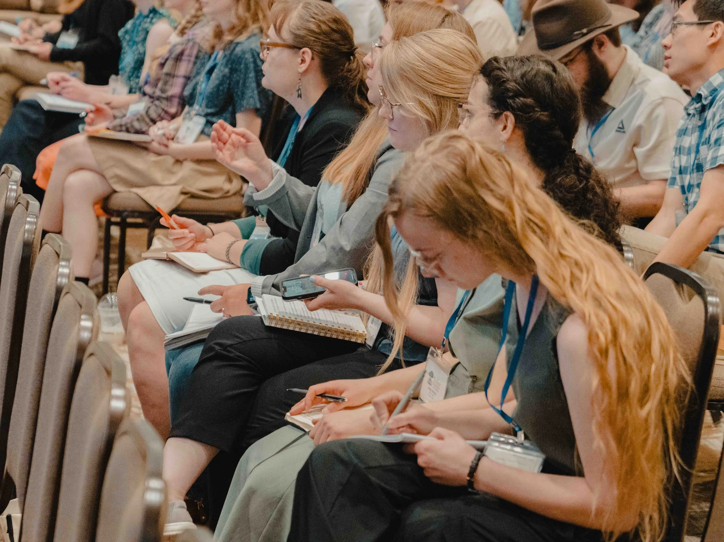 A group of people sitting in a conference or seminar, taking notes and listening attentively.