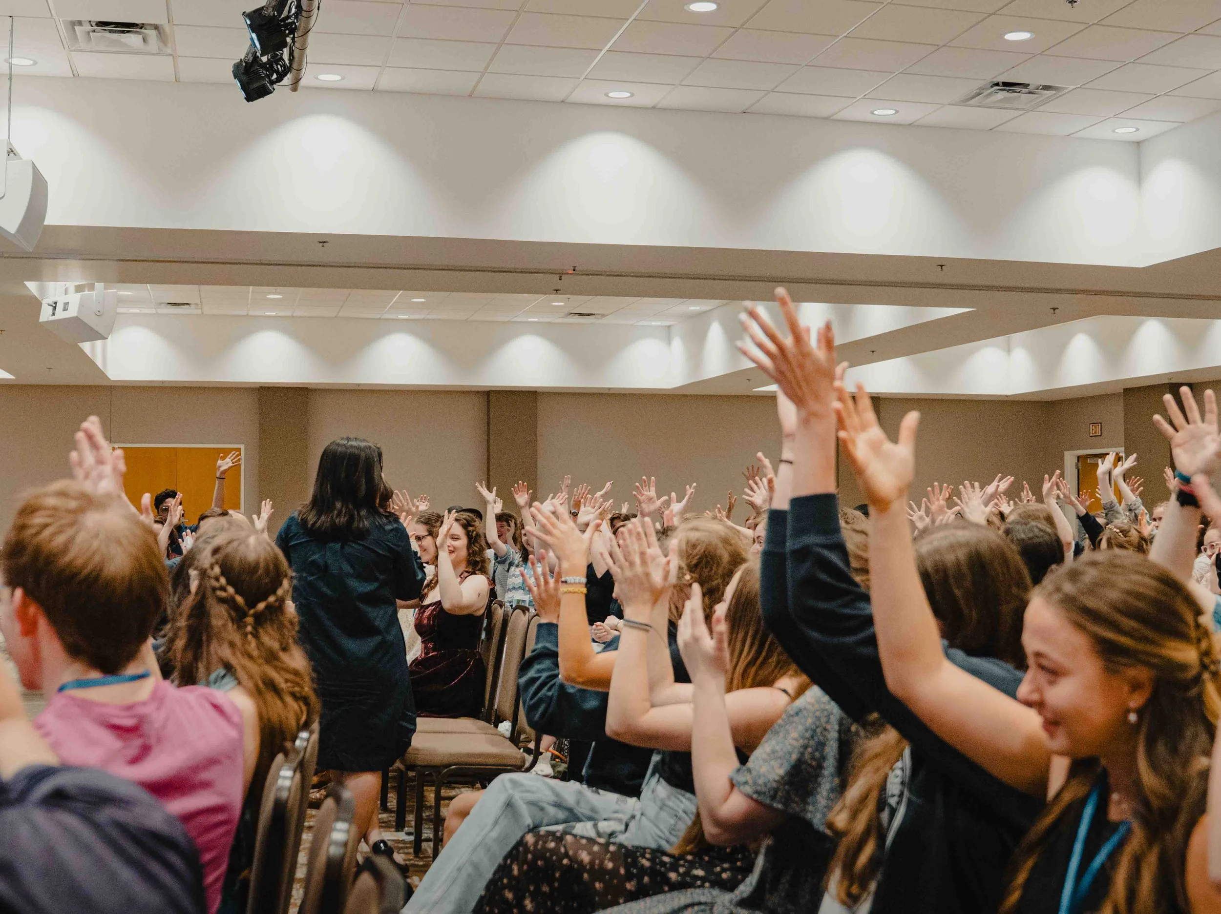 Many children and teenagers raising their hands in a large indoor room, possibly a classroom or assembly hall, with a high ceiling and bright lighting.