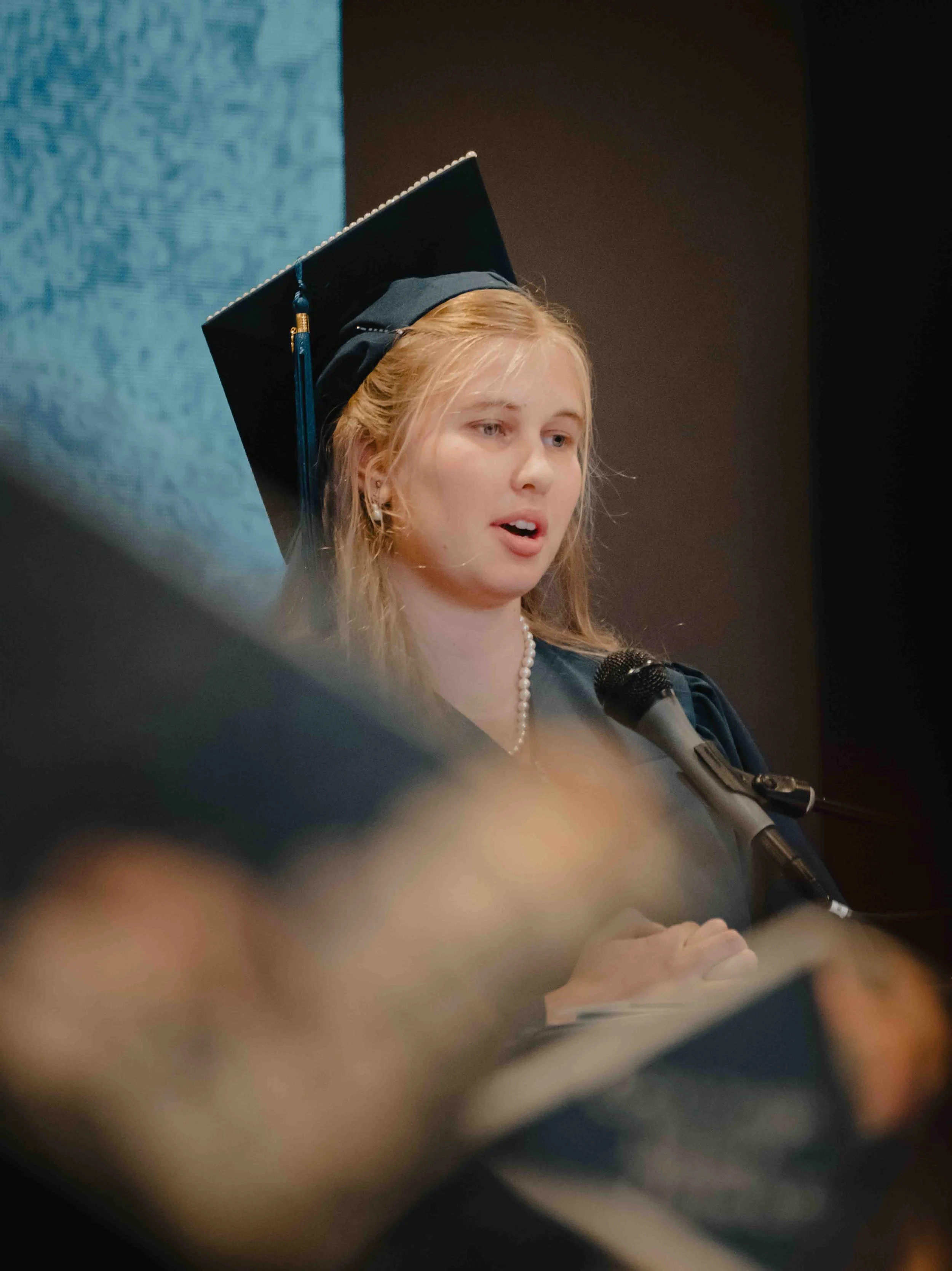 Young woman wearing a graduation cap and gown speaking into a microphone during a graduation ceremony.