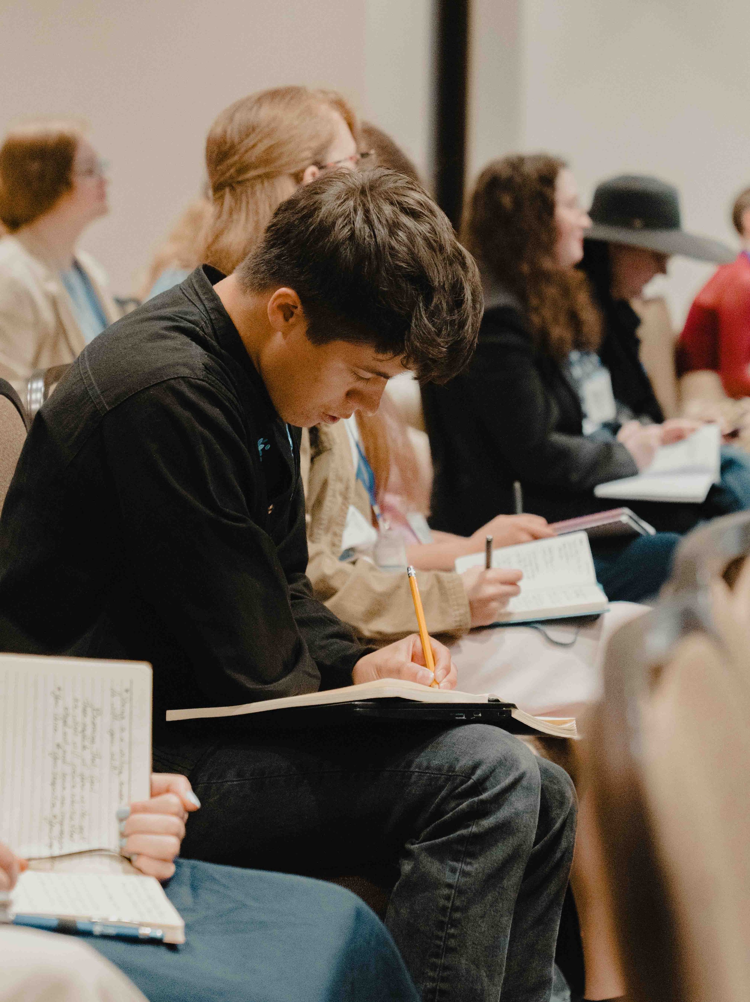 A group of people sitting in a row at a conference or seminar, taking notes and listening attentively.