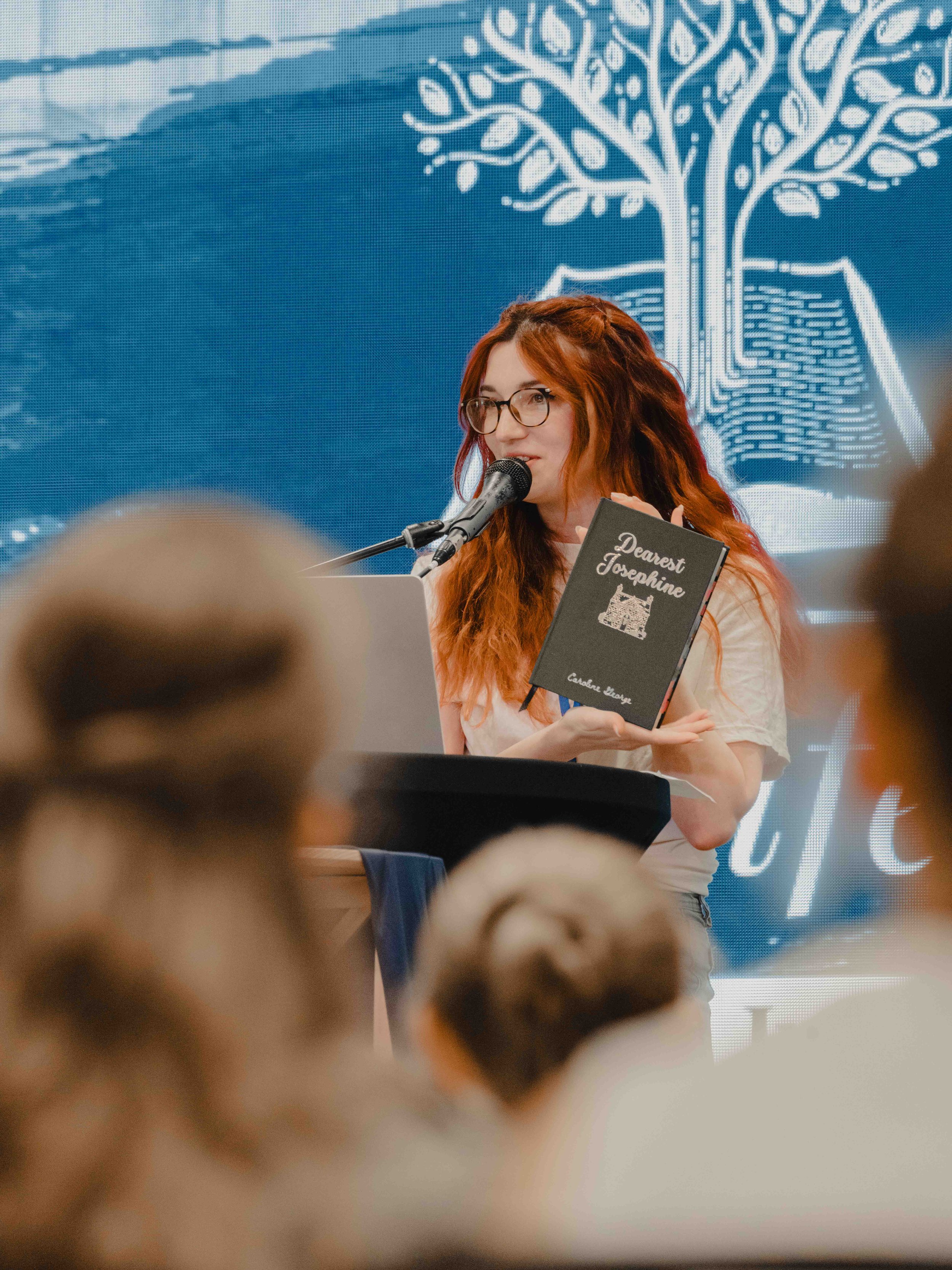 A woman with red hair and glasses is speaking into a microphone at an event, holding a black book titled 'Dearest Josephine.' In the background, a digital screen displays a white tree design.