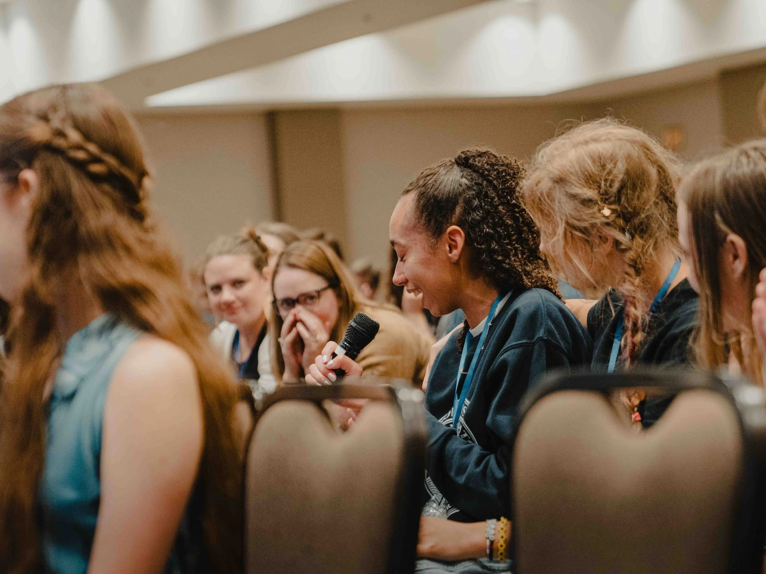 A group of women sitting in a conference or seminar, with one woman speaking into a microphone.