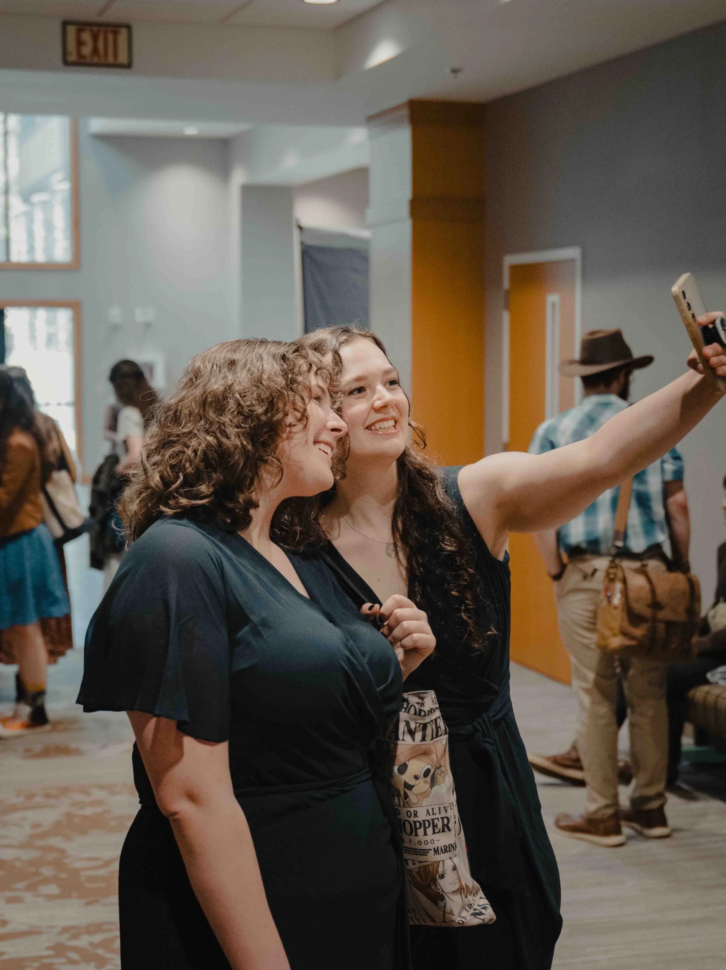 Two young women with curly hair taking a selfie together at an indoor event, with a crowd of people in the background.