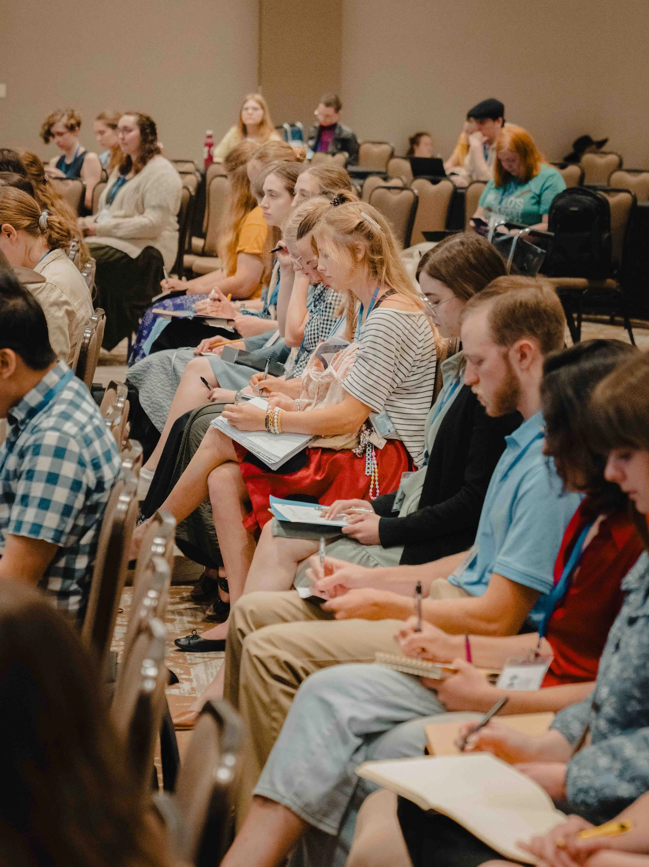 A group of young adults sitting in rows of chairs at an indoor conference, attentively taking notes and listening to a presentation.