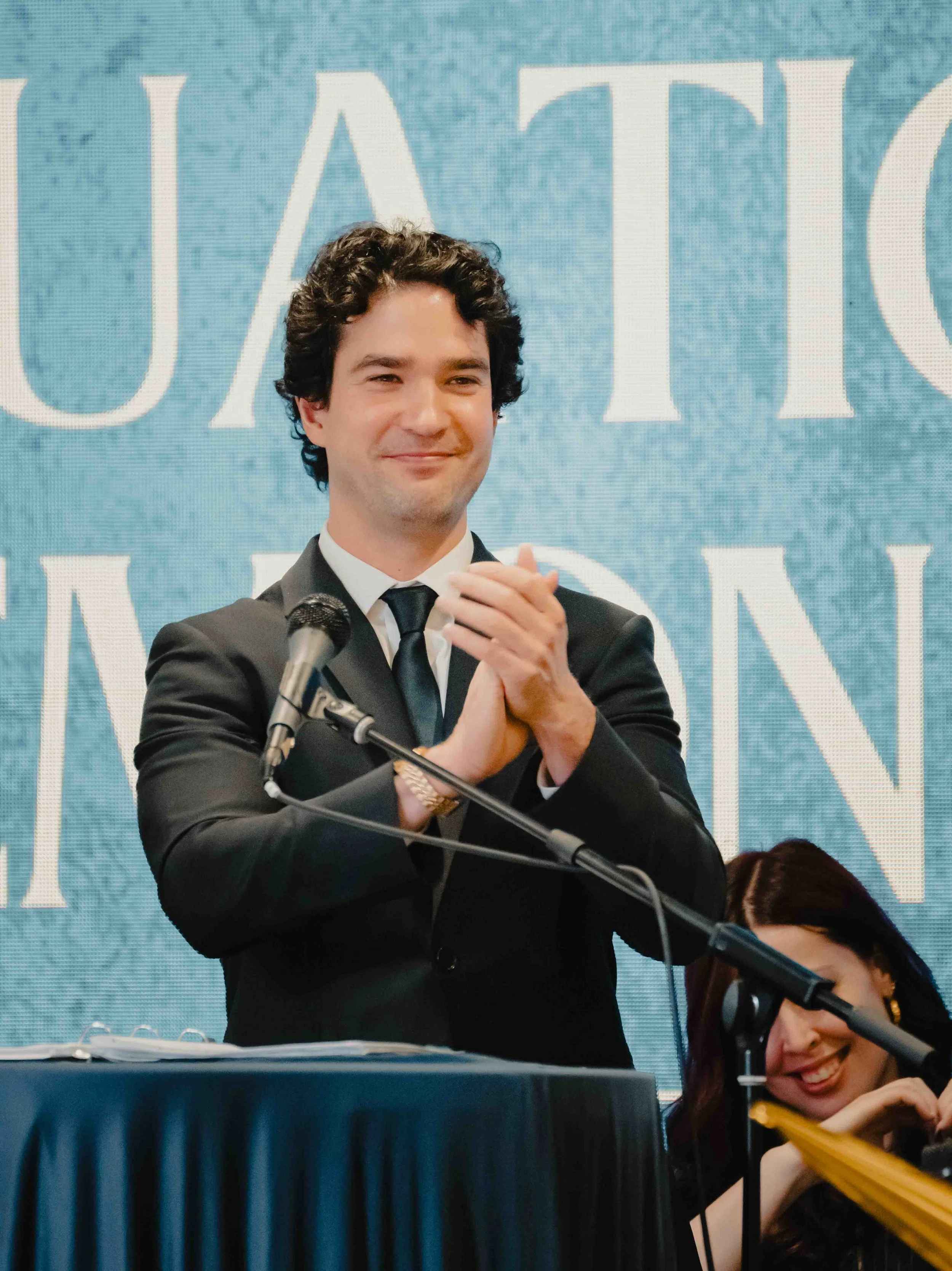 A man in a black suit and tie clapping and smiling at a conference or event behind a microphone, with a blue textured background and large white letters partially visible, with a woman with dark hair and glasses sitting and smiling at a desk in front