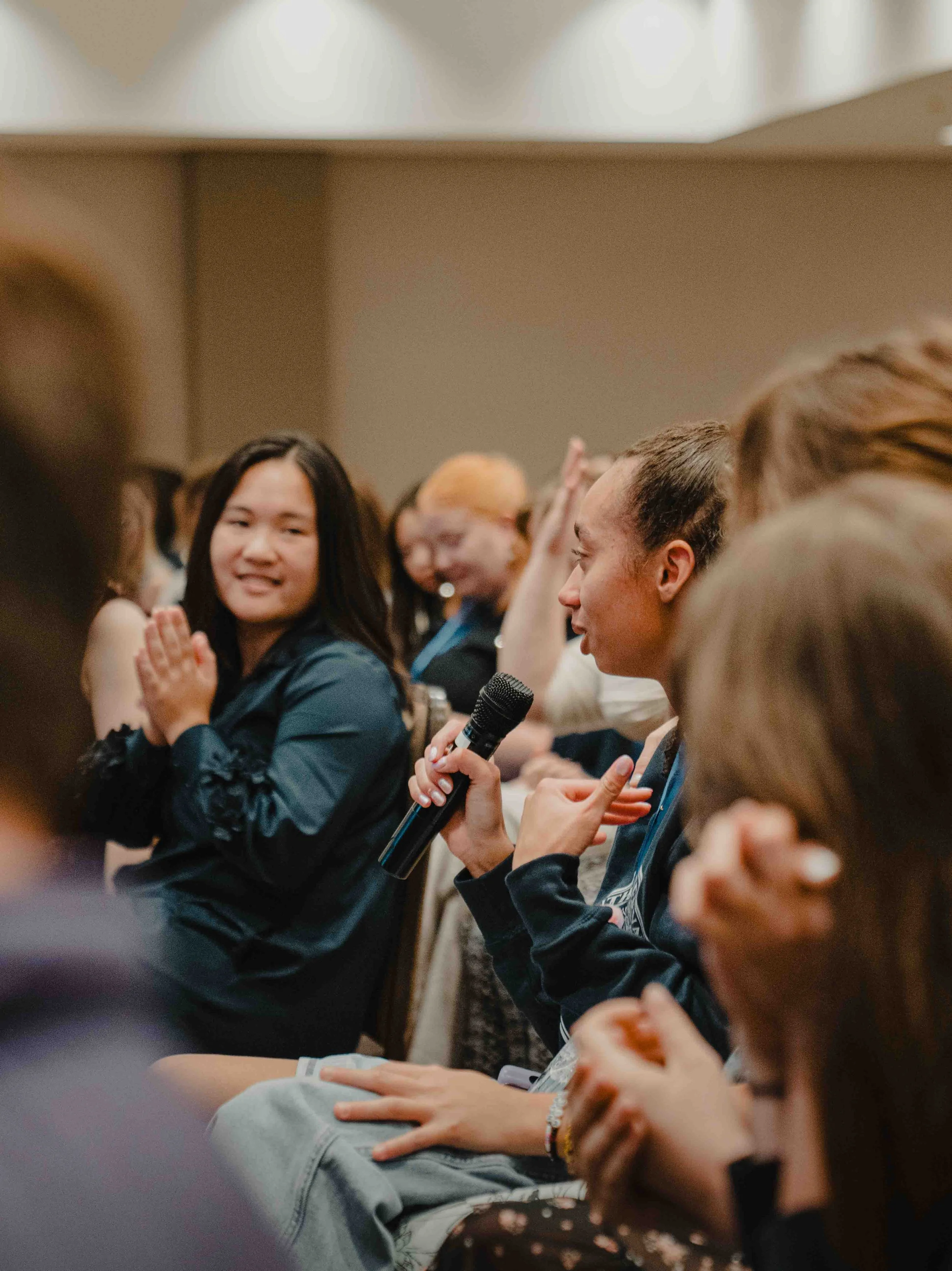 A diverse group of people sitting in a room, engaging in a discussion, with some holding microphones and others clapping, indicating a participatory event or conference.