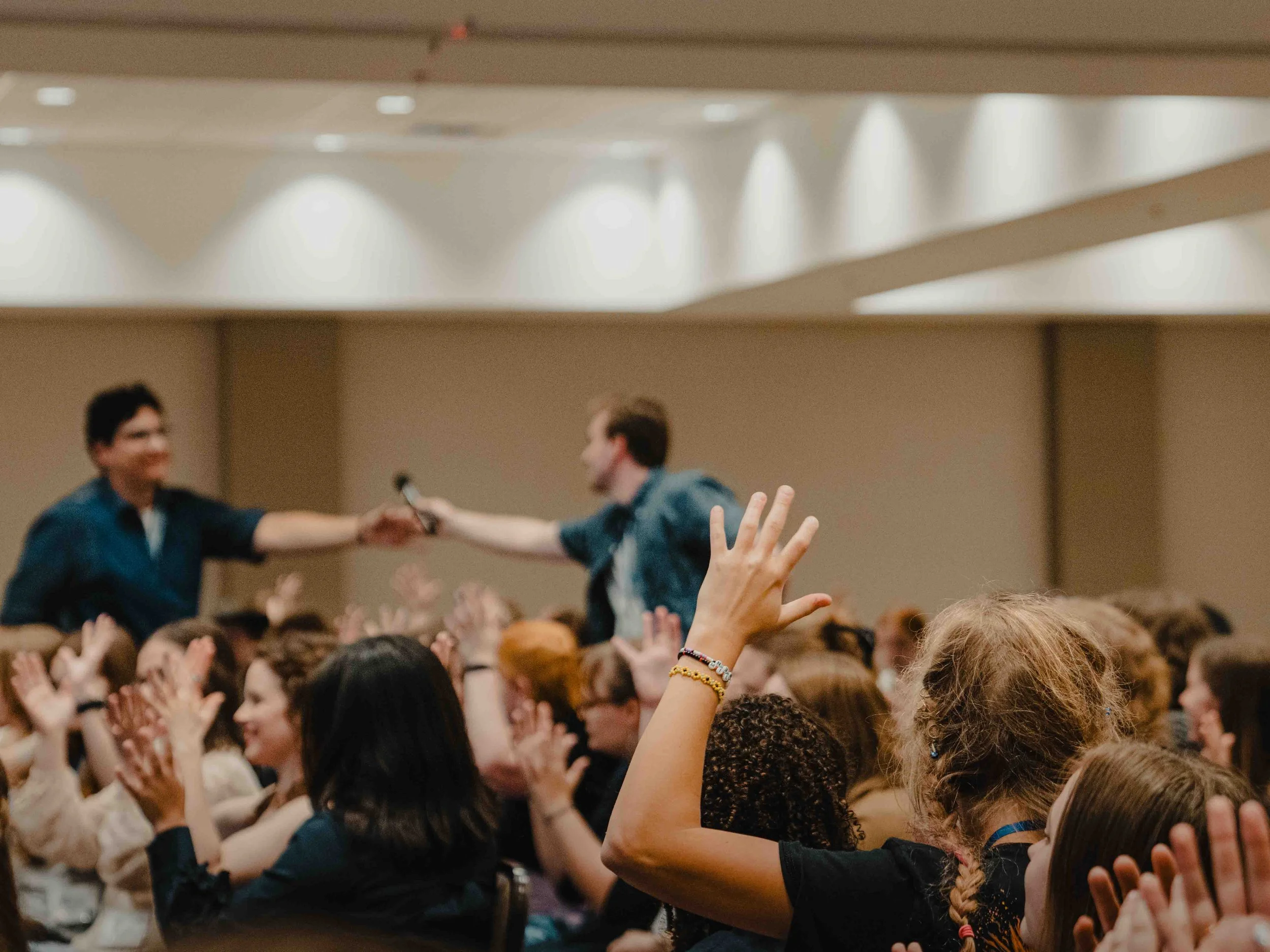 A large group of people seated in an auditorium with many raising their hands. Two individuals on stage are shaking hands and engaging with the audience.