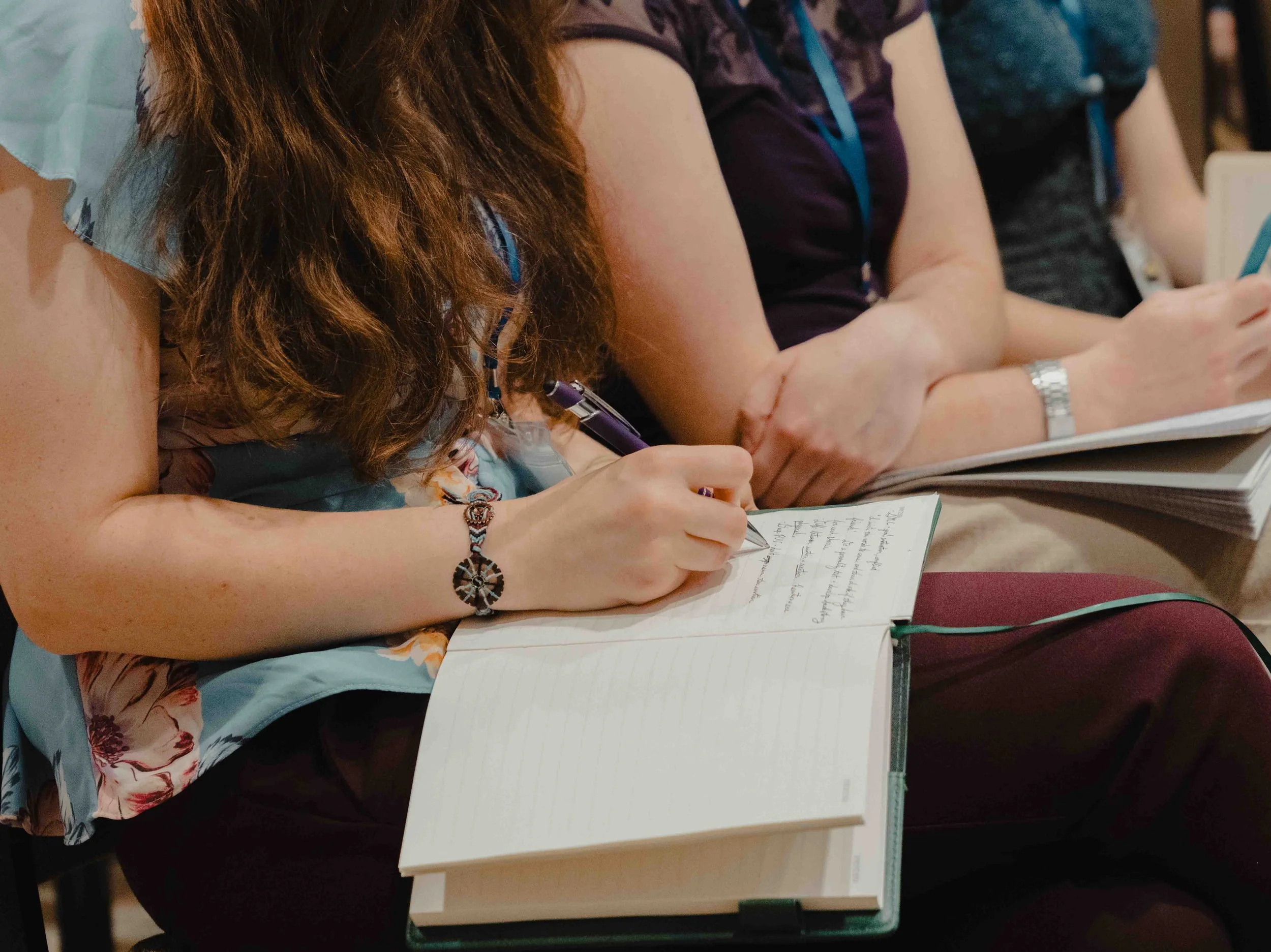People sitting at a table taking notes and using electronic devices during a conference or meeting.