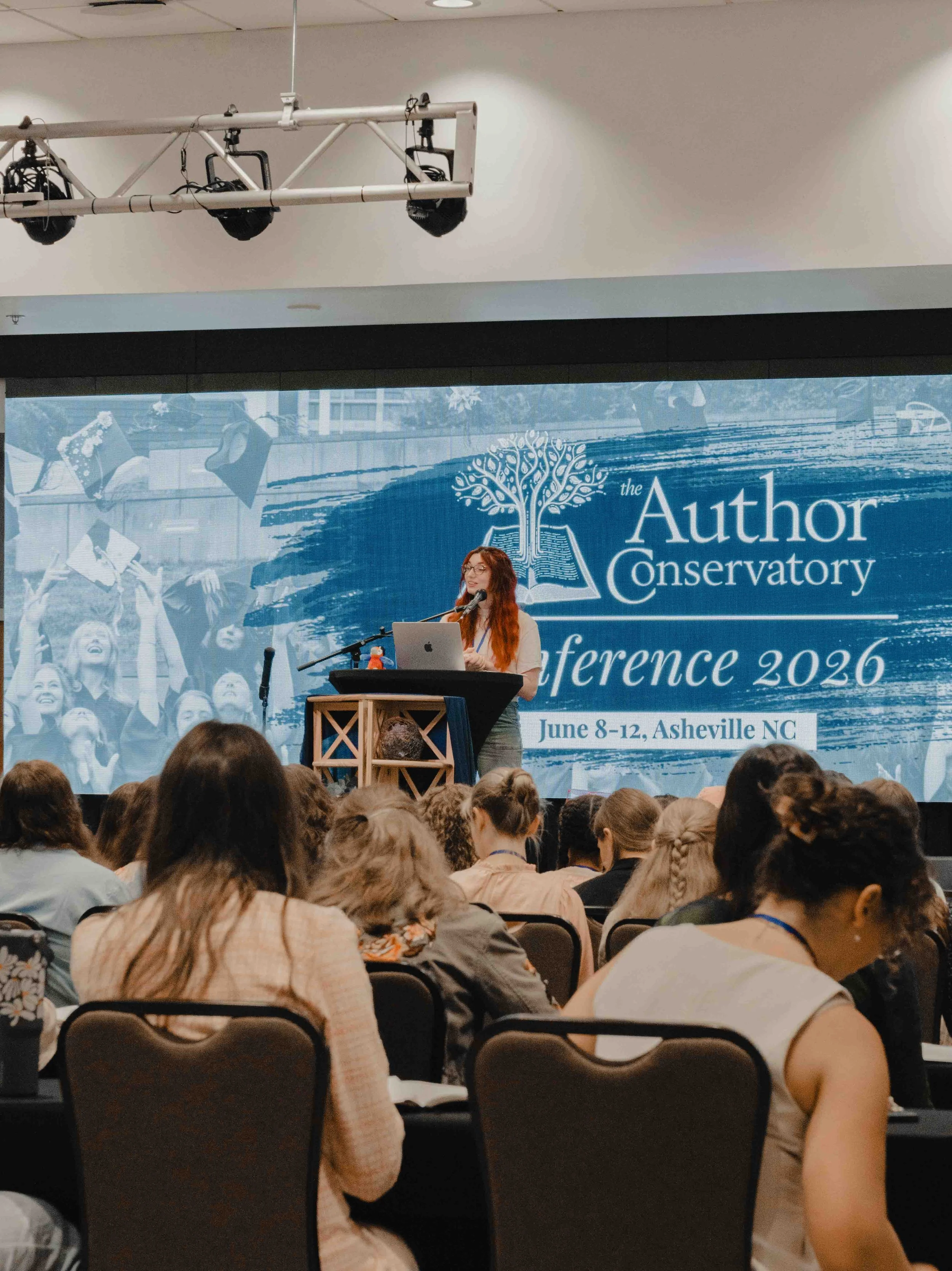 A woman with long red hair and glasses is standing at a podium with a microphone and a laptop, speaking at what is likely a conference. Behind her, a large blue banner displays the words "the Author Conservatory" and the dates June 8-12, Asheville NC