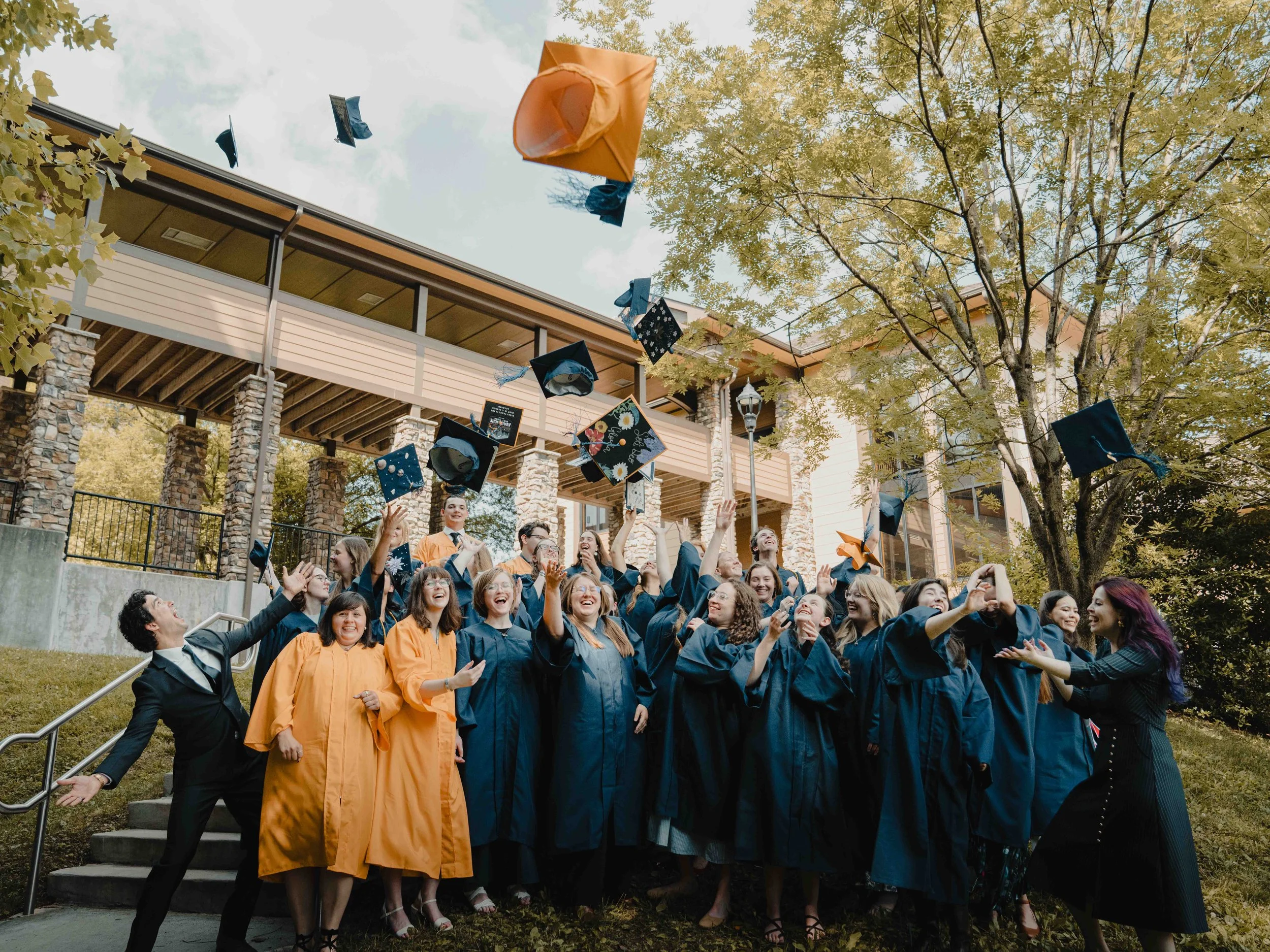 Group of young adults wearing graduation gowns celebrating outdoors, tossing caps in the air, with a building, trees, and cloudy sky in the background.