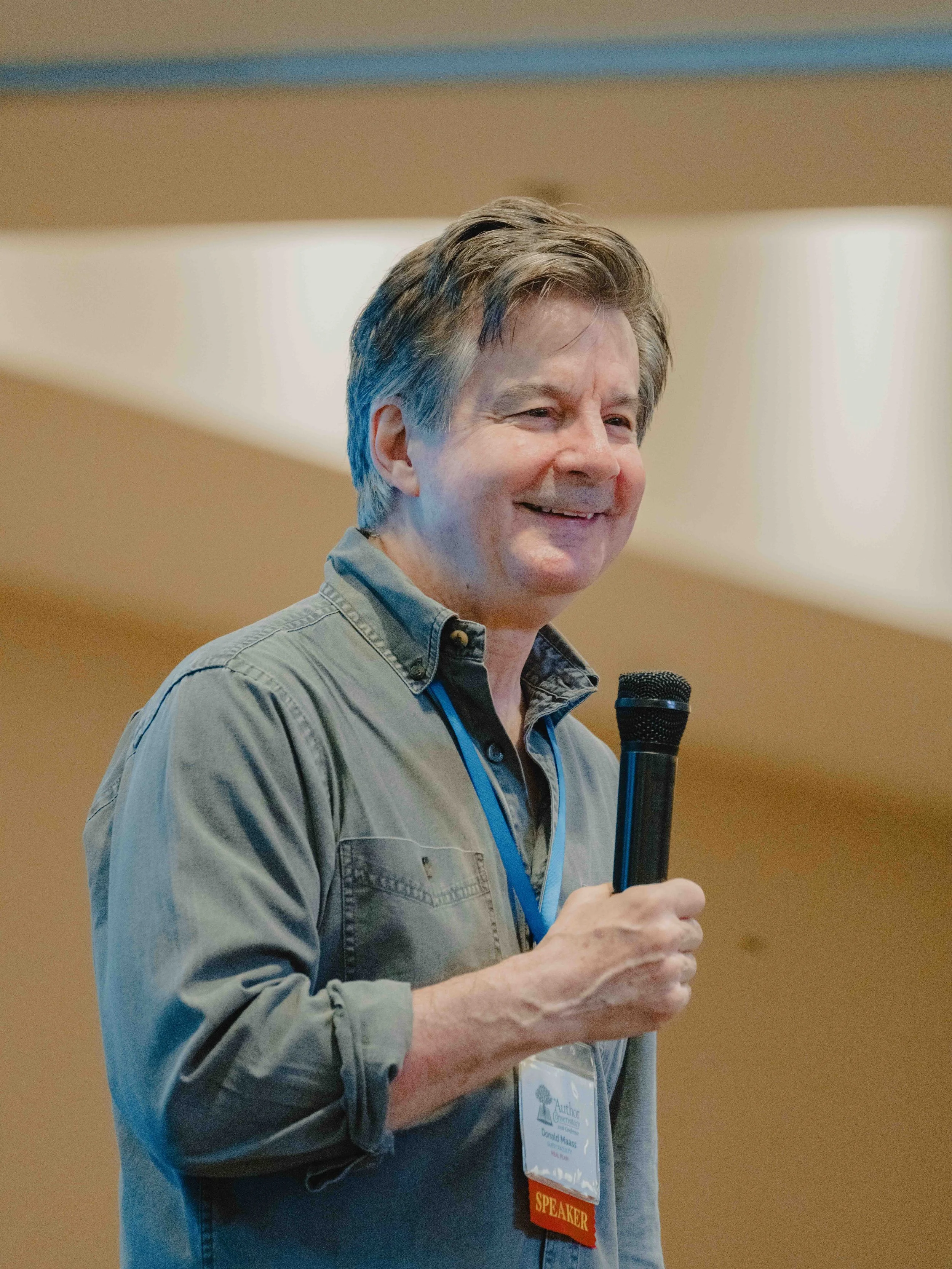 A middle-aged man with gray and brown hair smiling while holding a microphone. He is wearing a gray shirt with rolled-up sleeves and a conference badge around his neck. The background appears to be an indoor setting.