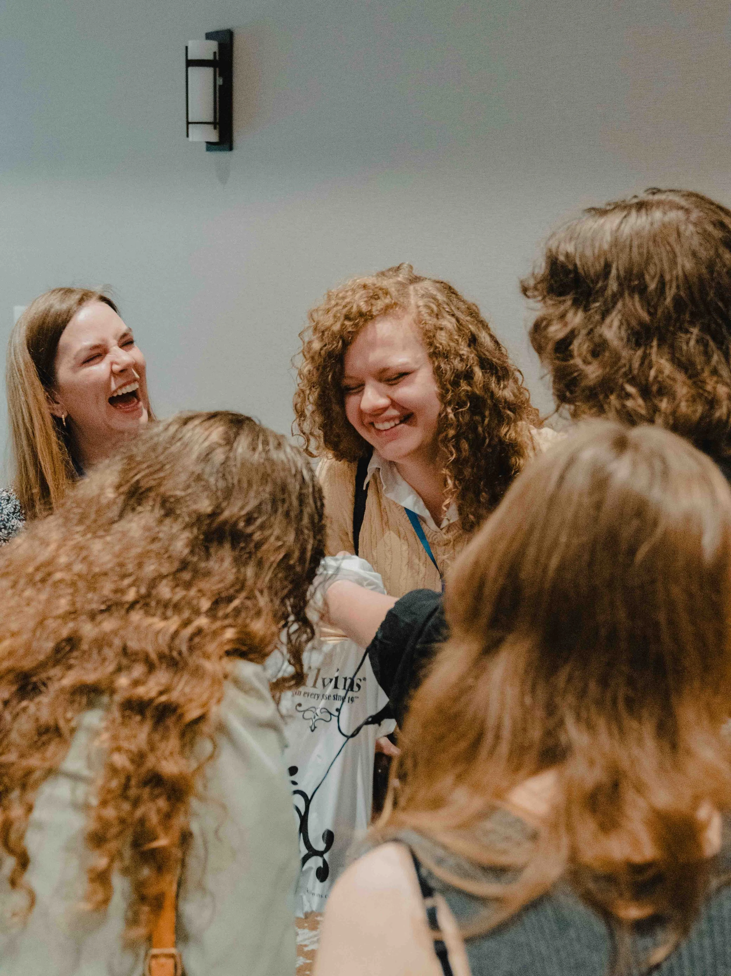 Group of people, mostly women, gathered in a circle, laughing and smiling, in an indoor setting with a plain gray wall in the background.
