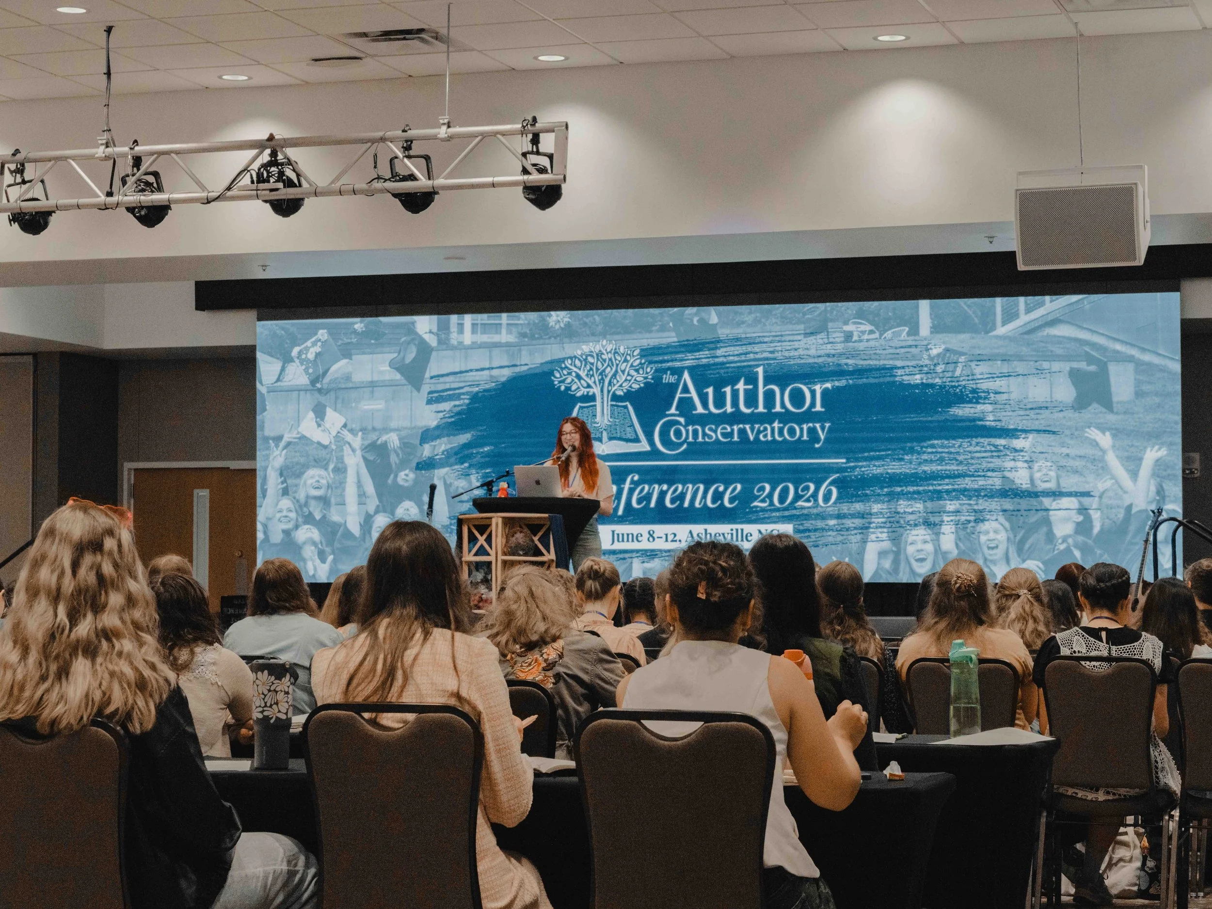 A woman with red hair and glasses is speaking at a conference stage with a large blue screen behind her. The screen displays the conference name 'the Author Conservatory Conference 2026' and the dates June 8-12 in Asheville, NC. The audience is seate