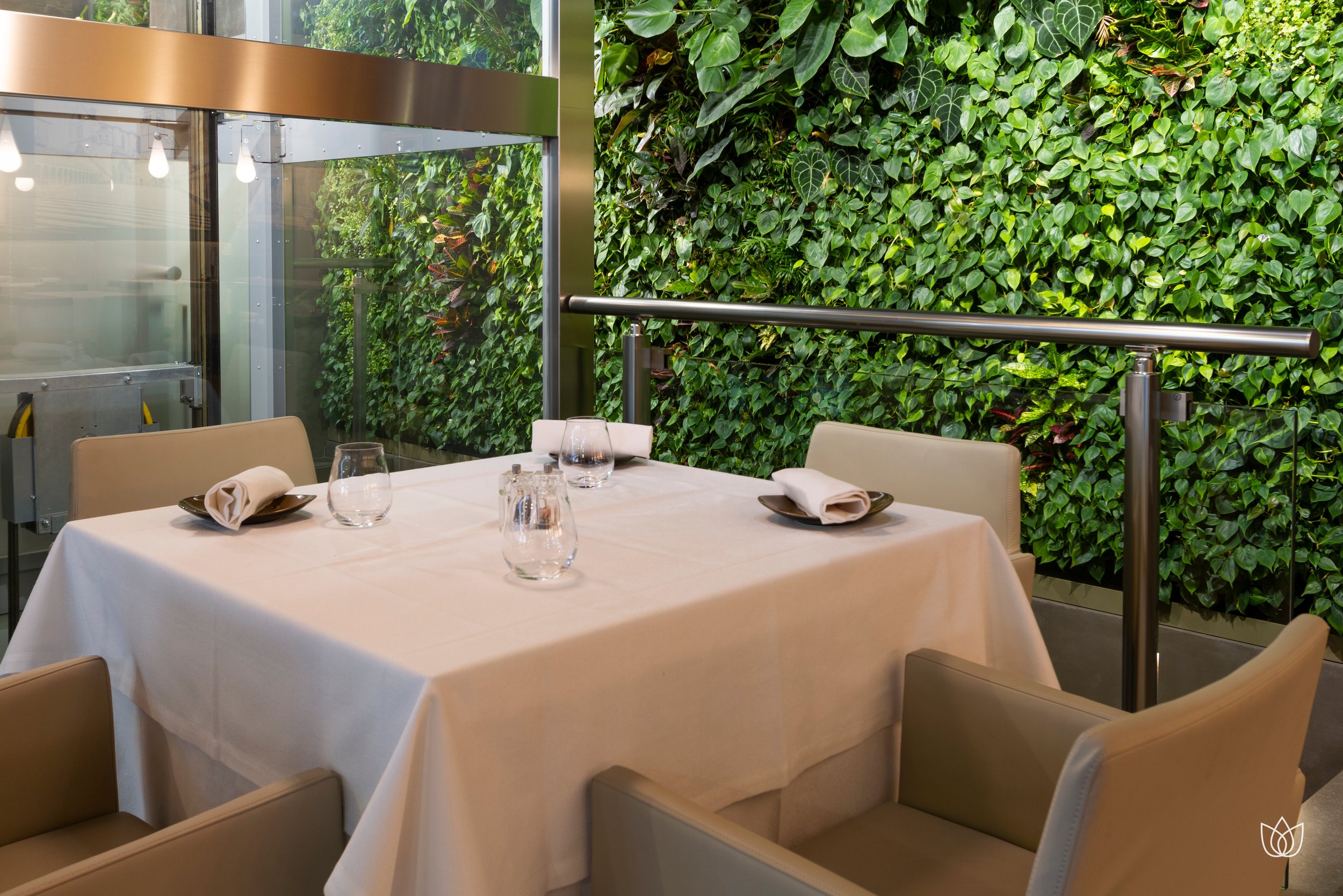Dining table with white tablecloth, cutlery, glasses and napkins, in a restaurant with a green wall of plants