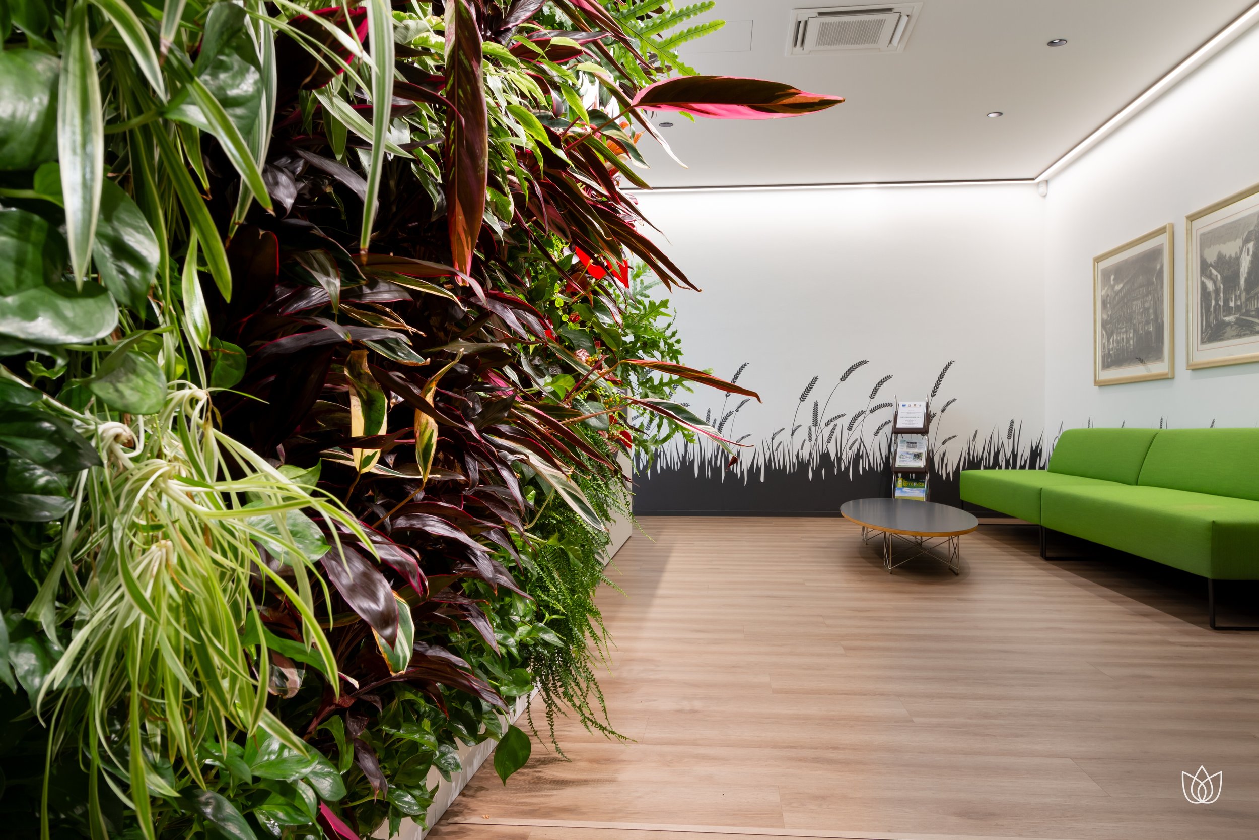 The lobby of a facility with a wall of green and red plants on the left, a green sofa on the right, and a small table in the center, with white walls decorated with drawings and two paintings, with a wooden floor.