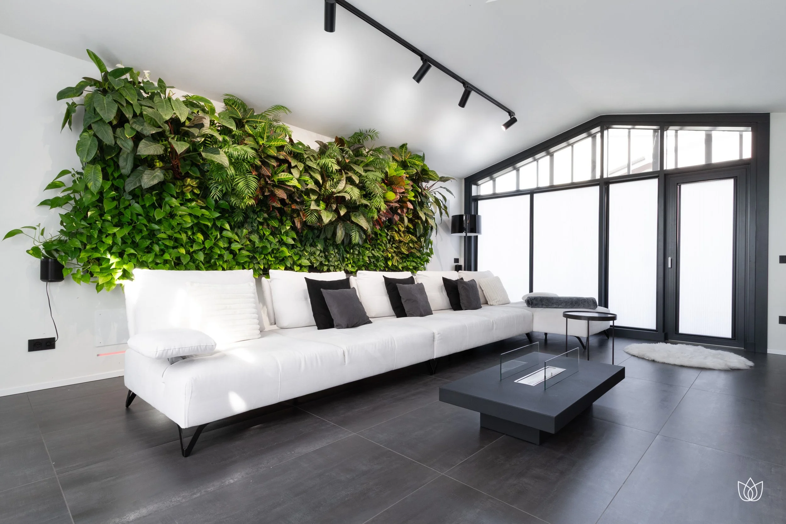 Modern living room with large white sofa, black and gray cushions, green wall with plants, large window with black frame, and sliding door.