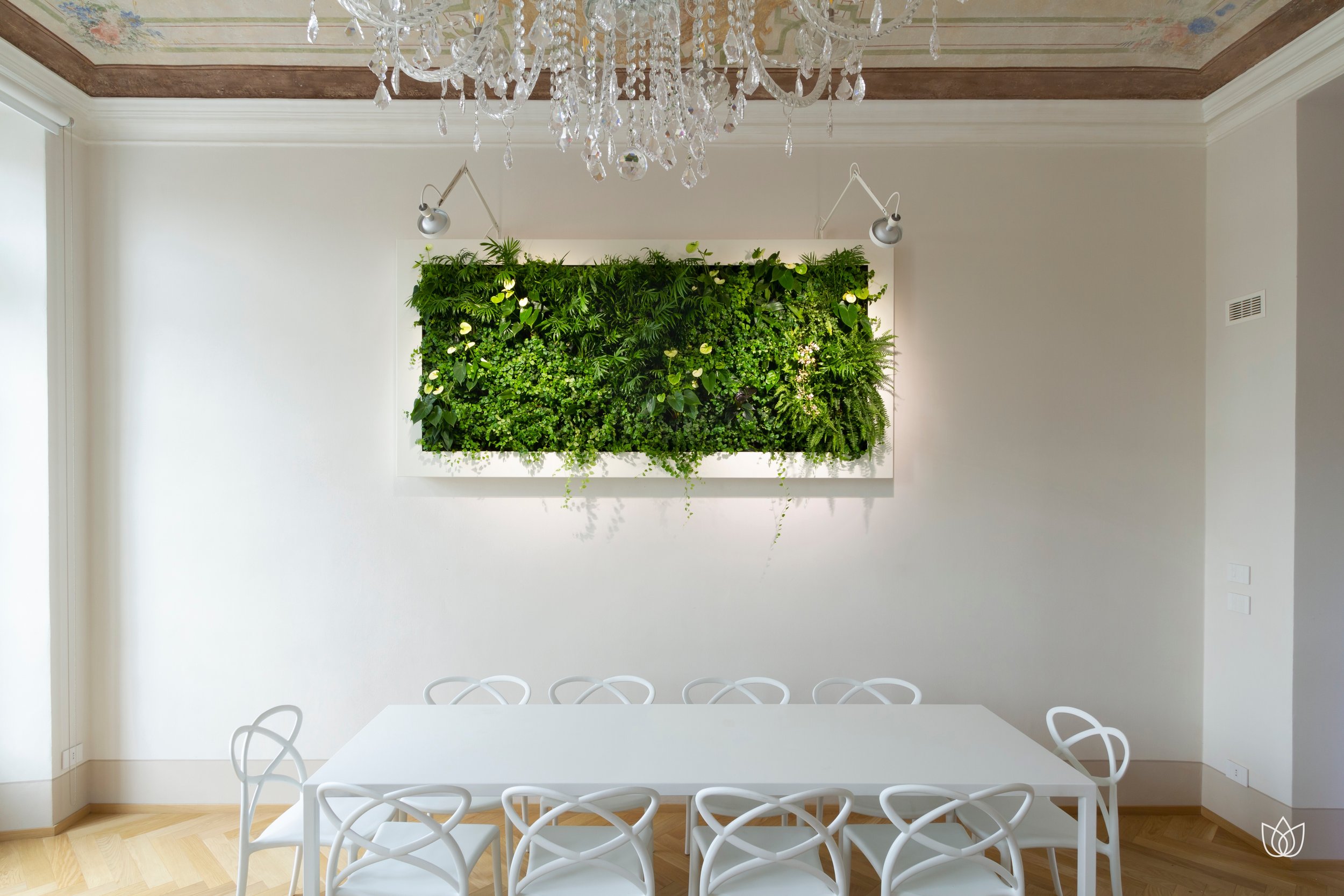 Dining room with white table and white chairs, green wall with plants and lamps above.