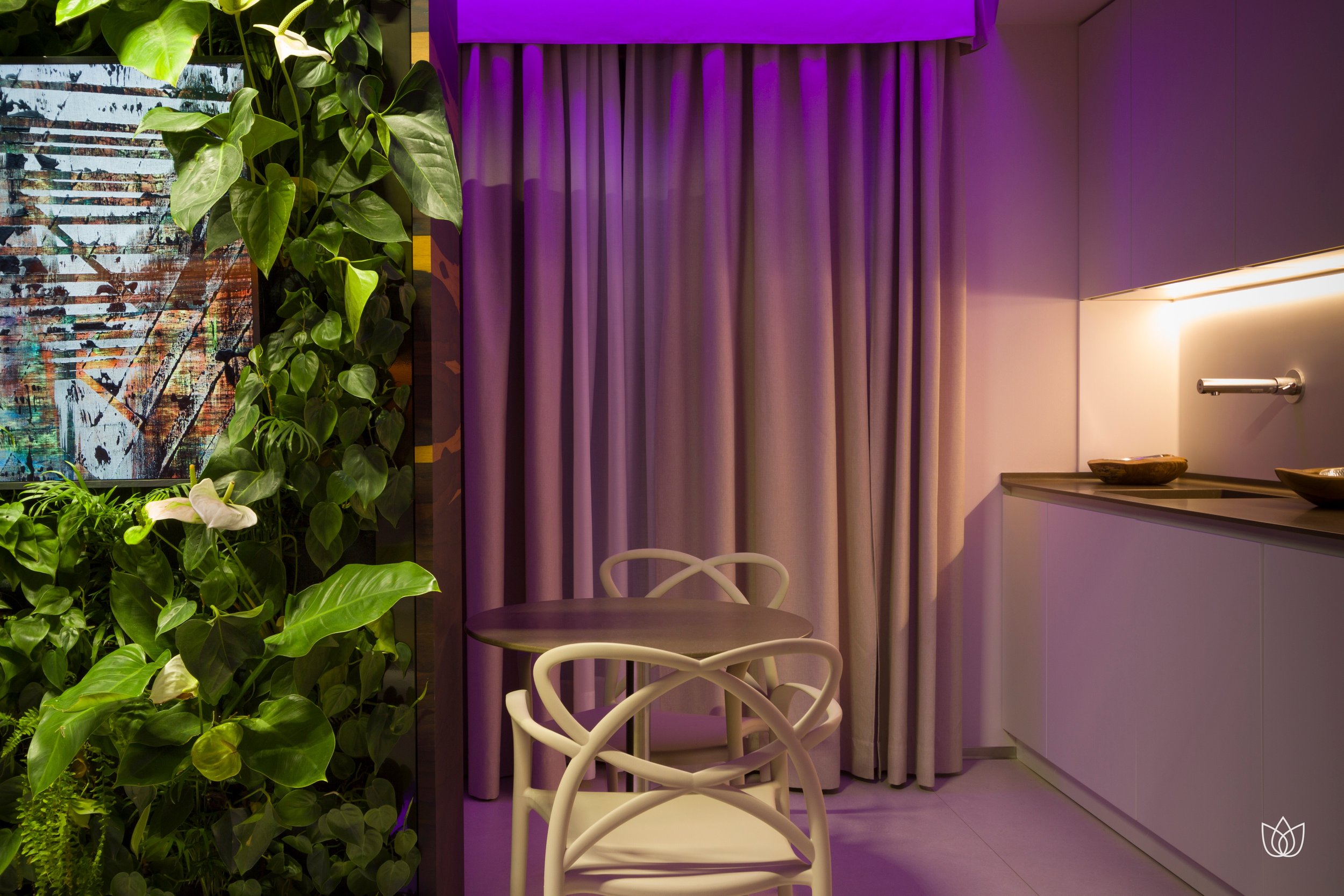 A corner of an interior with a wall of green plants, bare curtains, round coffee table and white chairs with soft purple lighting.