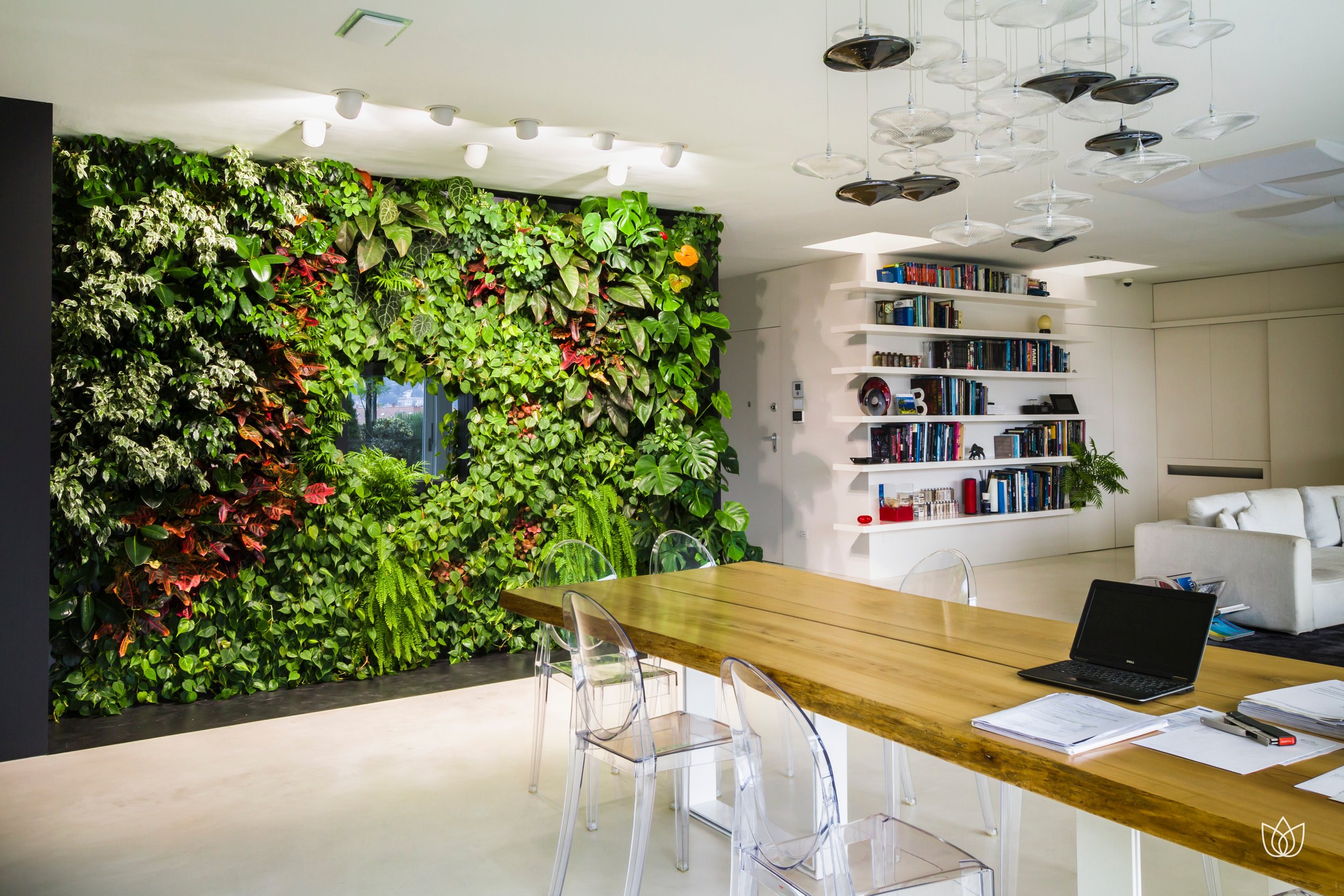 Modern room with green wall of plants and books, large wooden table with transparent chairs, white bookcase with objects and plants, white sofa.