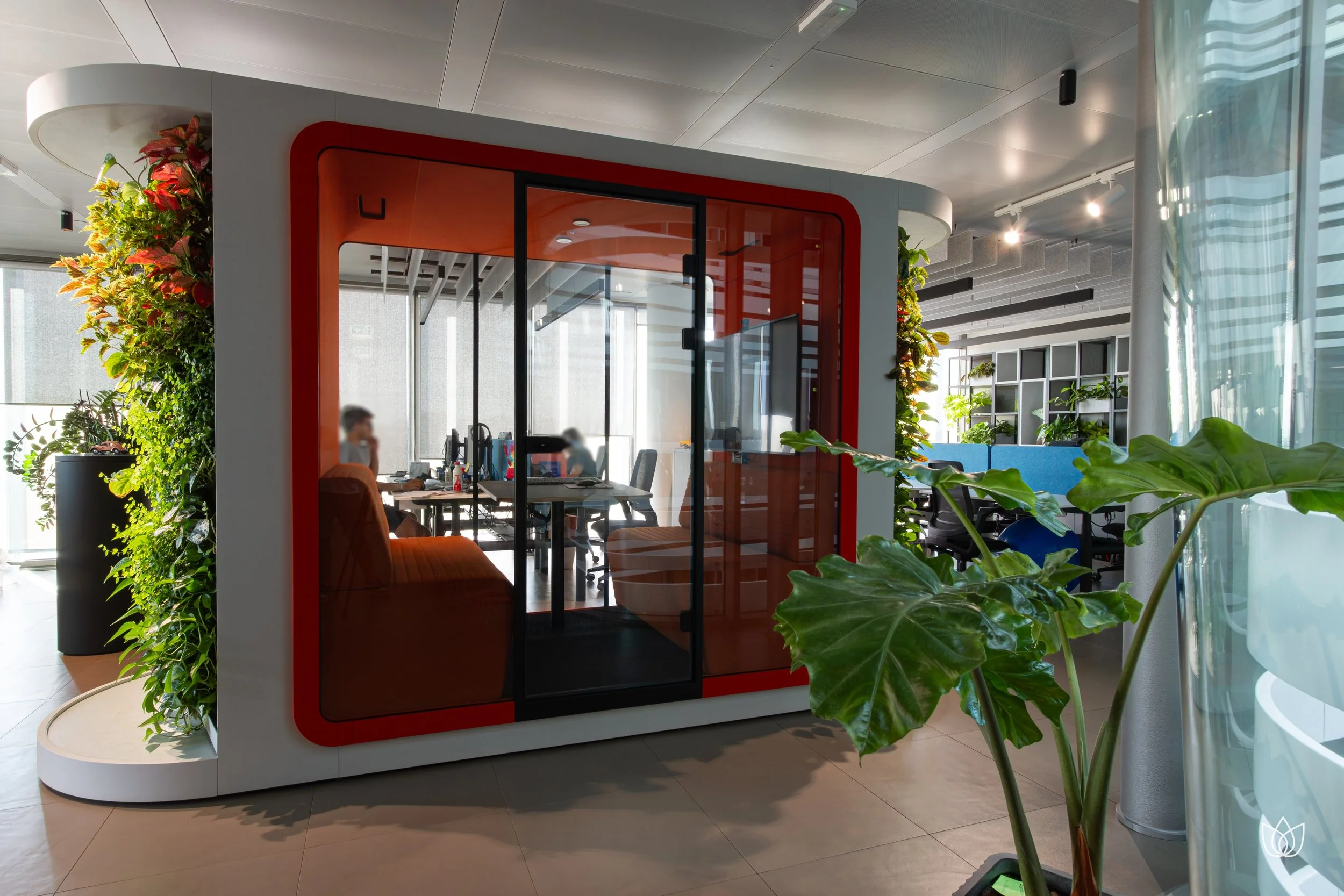 Ambiente di ufficio moderno con una cabina di vetro arancione, piante verdi e scrivanie con persone al lavoro.