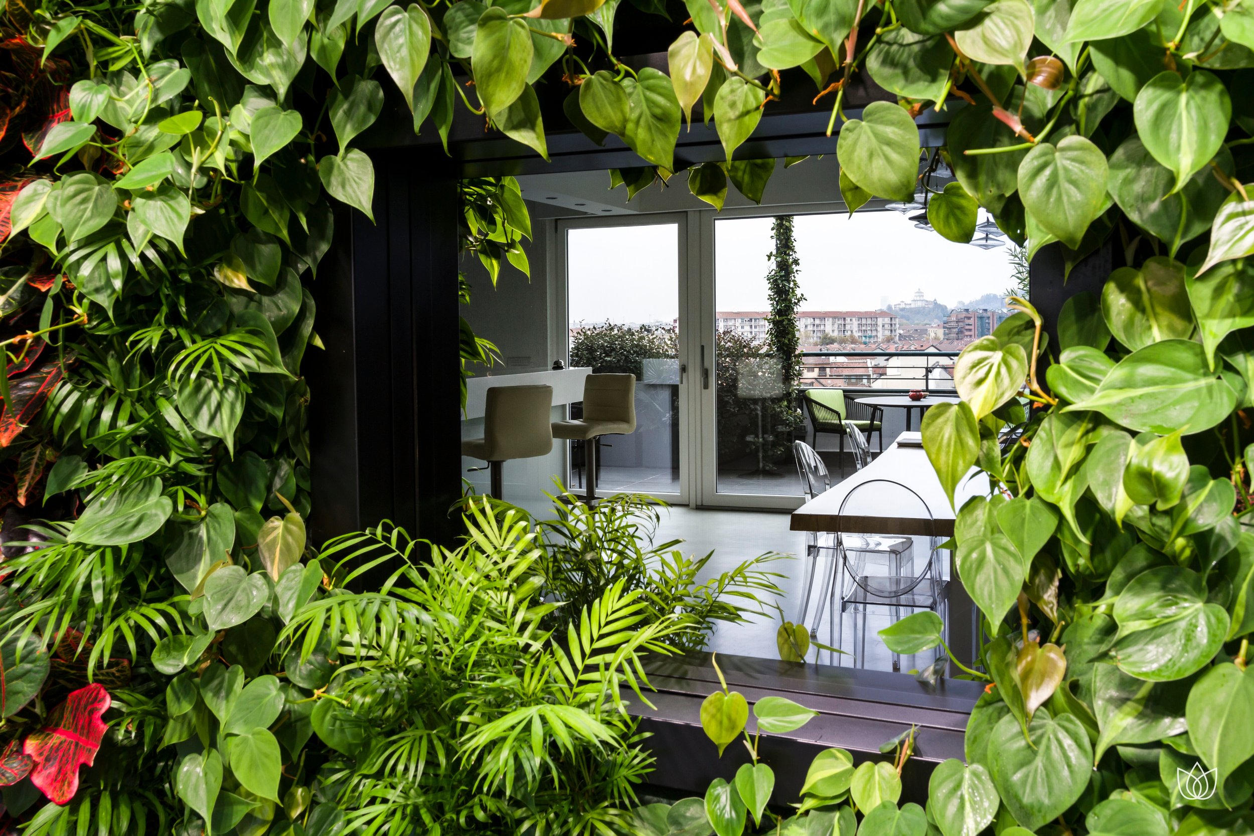 Interior of an apartment with a large window overlooking a balcony, decorated with many green plants that form an entrance or living area overlooking the city.