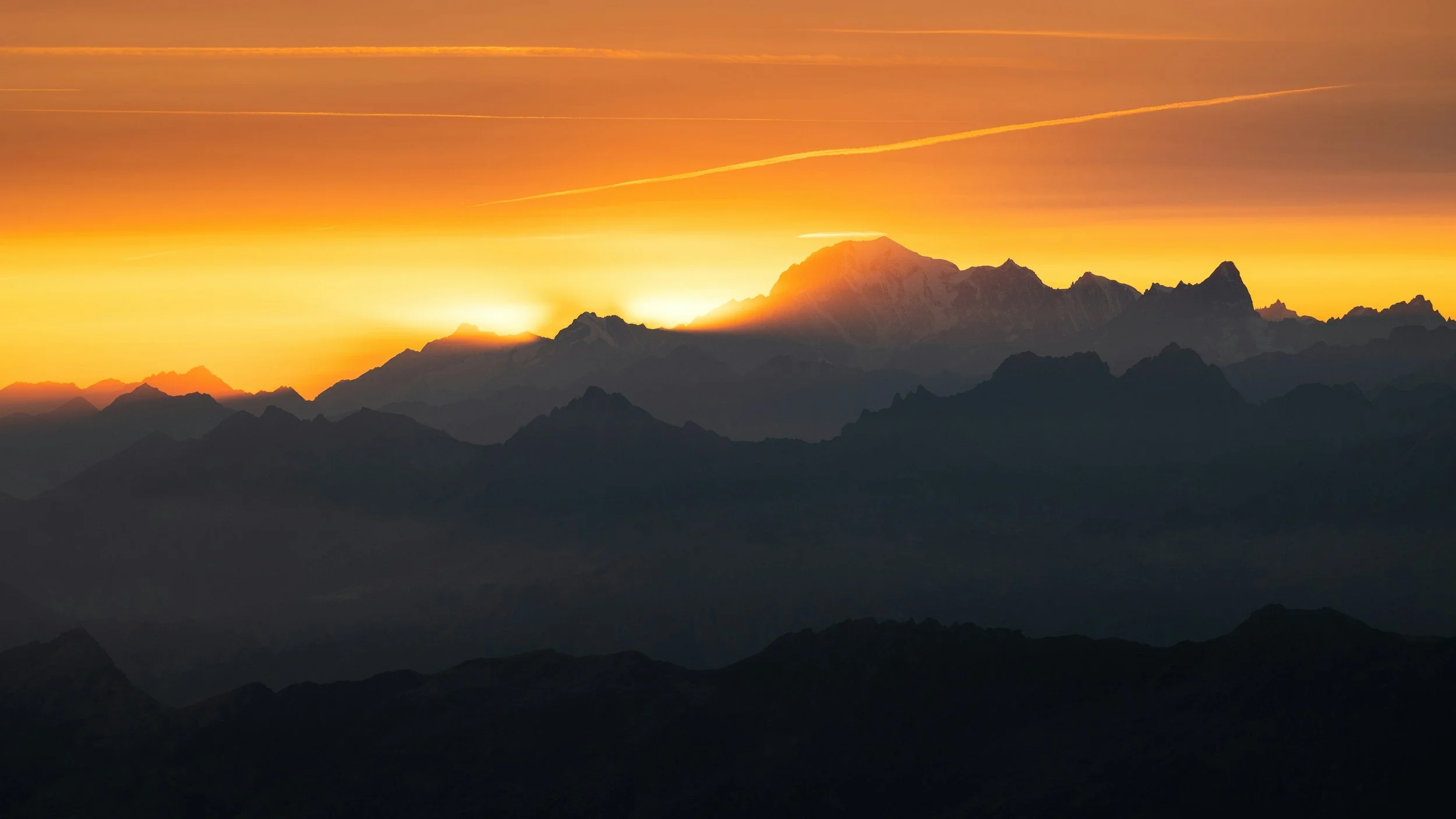 Sunset over mountain range with vibrant orange and yellow sky, silhouetted peaks, and distant clouds.