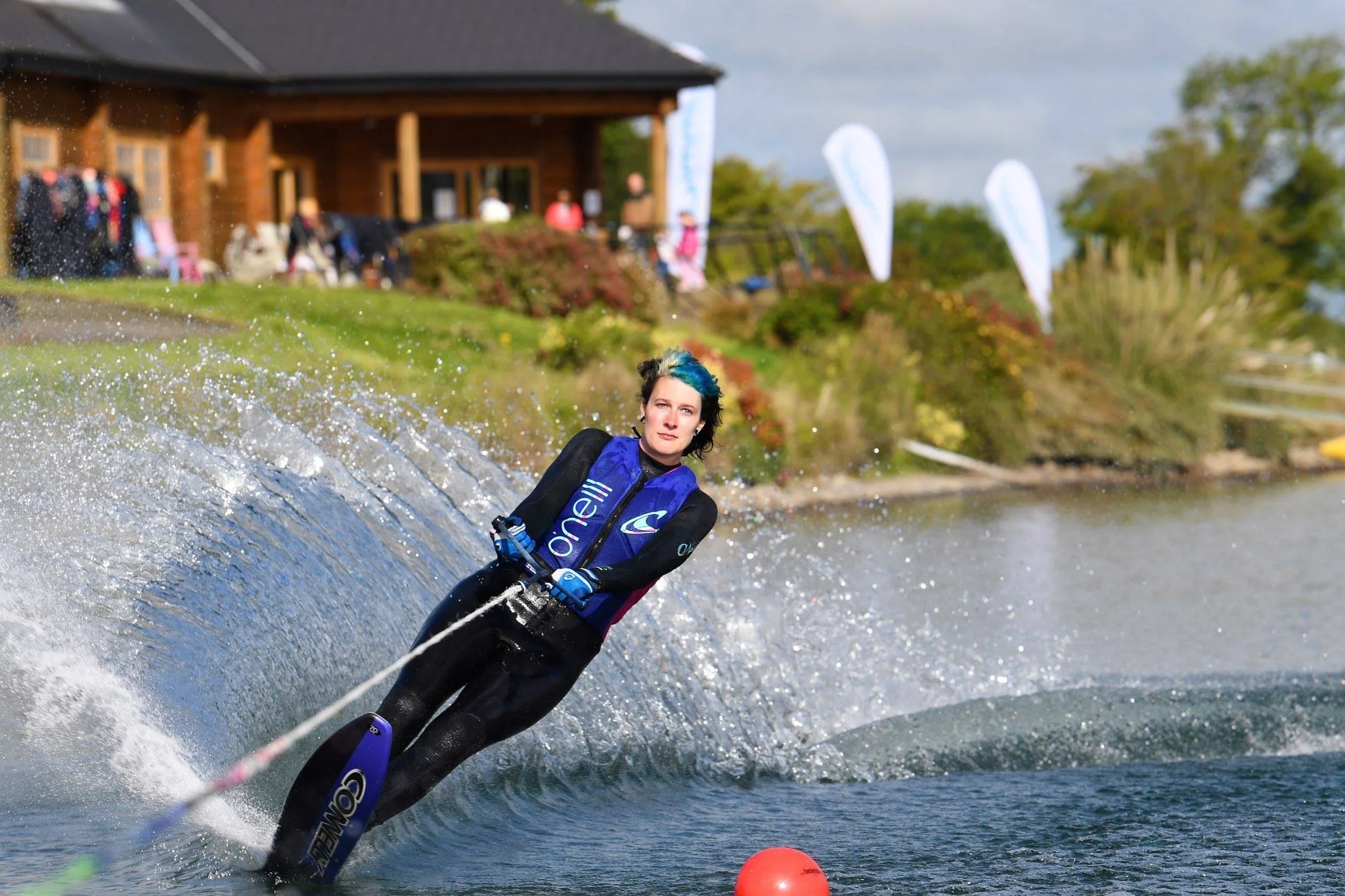 Person waterskiing on a lake, creating a spray of water, wearing a wetsuit with "O'Neill" logo, with a red buoy in the foreground and a building in the background.