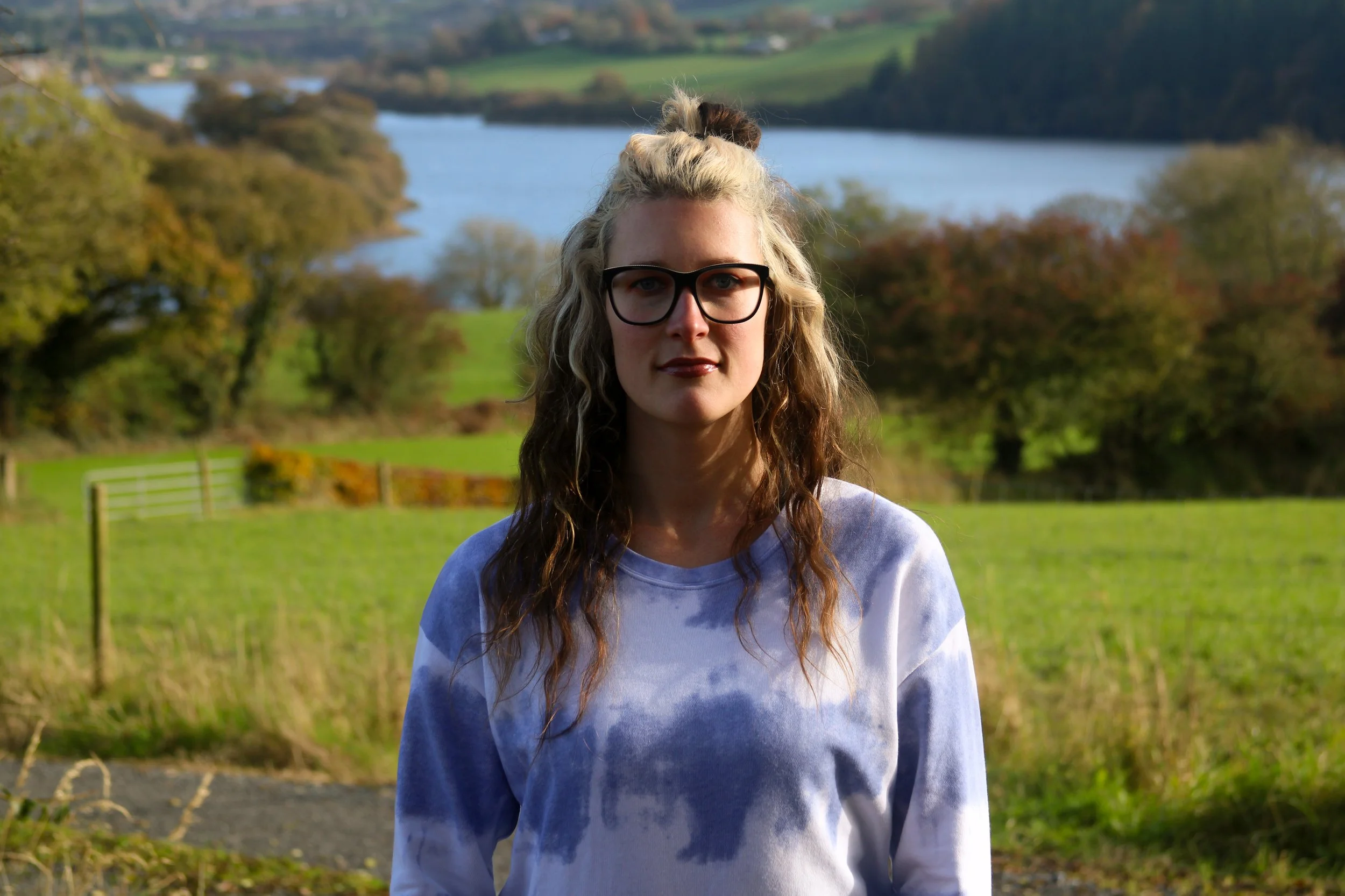 A young woman with blonde and brown wavy hair, glasses, and wearing a white and blue tie-dye sweater, standing outdoors in a green field with trees and a river in the background.